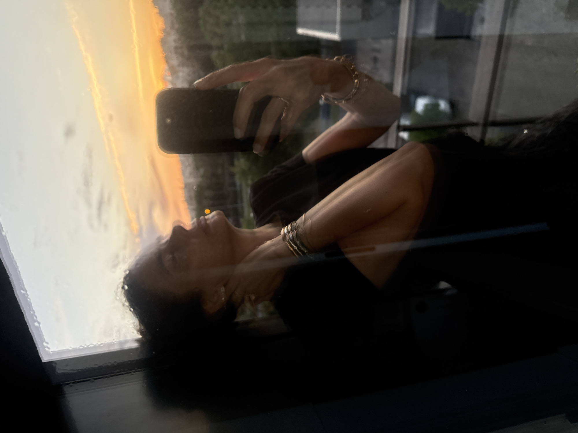 The image depicts a reflection of a person taking a selfie through a glass surface, possibly a window, at sunset or sunrise in Loures, Portugal. The main subject is a person, likely female, whose profile is visible along with her right arm. She has dark, curly hair and appears to be looking downwards or has her eyes closed, conveying a contemplative or serene expression.
Her right hand holds a dark smartphone, poised for taking a picture. She wears rings on her fingers and multiple bracelets or bangles on her right wrist. The glass surface she is reflected in is covered with numerous small water droplets, suggesting rain or high humidity.
Beyond the reflection of the person, the outdoor scene is dominated by a vibrant sky, painted in brilliant orange and yellow hues characteristic of a low sun angle. This warm, golden light suggests either dawn or dusk. In the deeper background, behind the person's reflection, indistinct shapes of buildings or other structures are faintly visible, indicating an urban or residential setting. The overall impression is one of quiet self-capture against a striking natural backdrop. There is no visible text in the image.