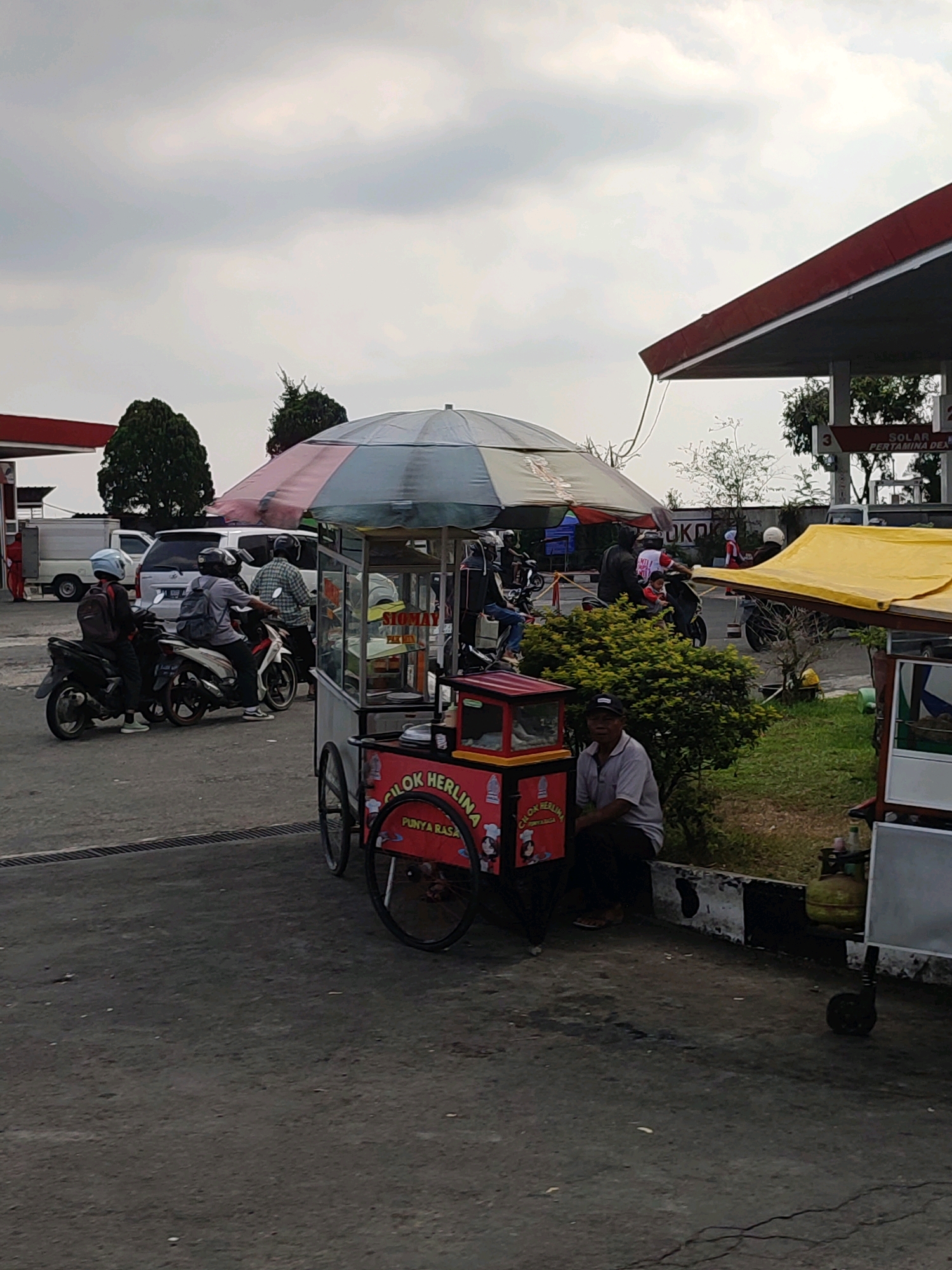 This image captures a street scene, likely at a gas station, in Bojasari, Indonesia. In the foreground, a man in a gray shirt and black cap sits on a low concrete ledge, facing the camera. To his left, a red food cart with a striped umbrella is parked. The cart displays text that appears to read "CILOK HERLINA" and "PUNYA RASA". To the right of the man, another food cart with a yellow canopy is visible, with a green gas cylinder on the ground next to it.
In the mid-ground, several people on motorcycles are present. One person in a blue helmet rides a black motorcycle, and another person in a gray shirt and black backpack is on a white motorcycle. Behind them, more motorcycles and people are visible, suggesting activity around the food vendors or the gas station. Various vehicles, including a white van and part of a white truck, are parked in the background.
The environment appears to be an outdoor paved area, possibly a parking lot or the grounds of a gas station. A red-roofed structure, likely part of a gas station canopy, extends from the right side of the frame, with signage indicating "SOLAR PERTAMINA DEX". Lush green bushes are present between the food carts and the gas station structure. The sky is overcast with visible clouds.