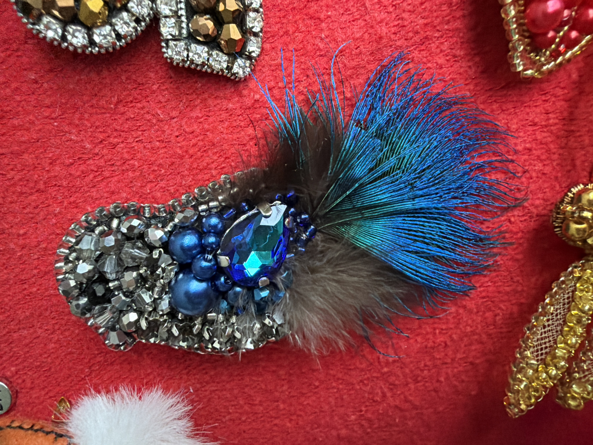 The image shows a close-up of a handcrafted brooch or embellishment on a red, textured surface. The brooch features a mix of materials including silver-toned beads, blue beads of varying sizes, a large, faceted blue jewel, and an arrangement of dark and iridescent blue feathers. The brooch's base is formed by closely packed silver beads, supporting the cluster of blue beads and jewel. A spray of feathers in shades of brown and blue fans out from behind the jewel. The textured red background suggests fabric or a similar surface. Fragments of other beaded adornments appear in the surrounding area, indicating that this brooch is among other similar items.