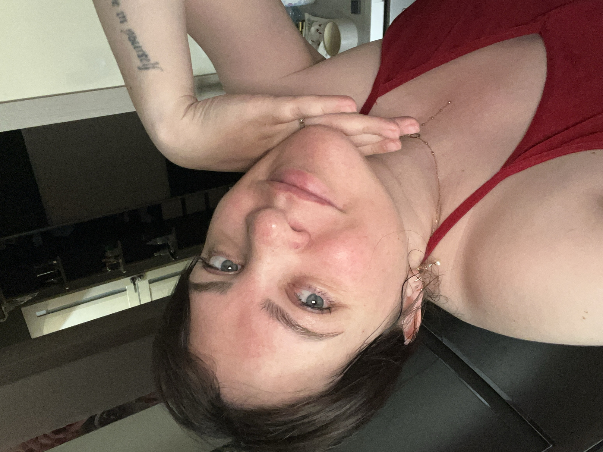 A woman with short brown hair, fair skin, and blue eyes is pictured in a close-up, angled selfie. She is wearing a red top and a delicate necklace. Her right arm is bent, with her hand resting on her chin, and she is wearing a ring on her right hand. A tattoo is visible on her right forearm, with text that appears to read "be in yourself". The background is dimly lit and appears to be an indoor domestic setting, possibly a closet or storage area, with shelves visible. The overall lighting suggests an indoor environment, potentially during nighttime or in a dimly lit room. There are no identifiable landmarks or objects that would definitively place the image in Noviy, Russia.