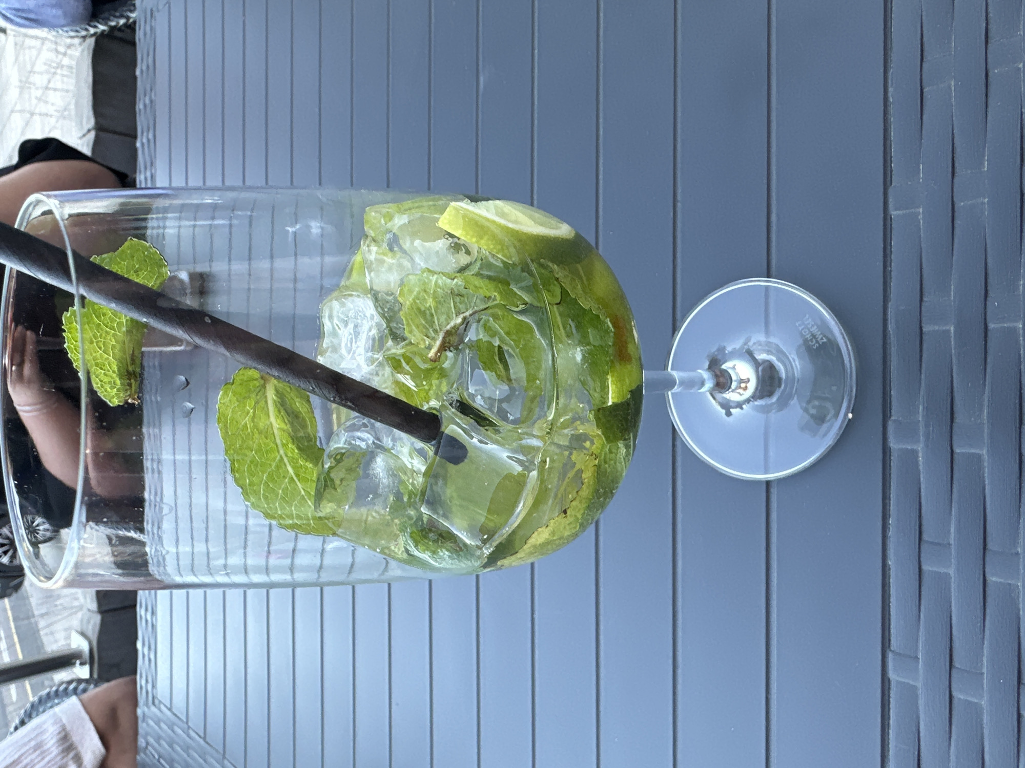 The image, taken in Käerjeng, Luxembourg, displays a refreshing drink in a tall, clear glass, likely a pitcher or large tumbler, set on a grey, slatted outdoor table. The glass is filled with numerous ice cubes, fresh green mint leaves, and slices of lime, strongly suggesting a mojito or a similar mint-lime beverage. A dark-colored straw is inserted into the drink.
To the right of the main glass, a clear, empty stemmed glass, resembling a wine glass, stands on the table. Visible text on the base of this stemmed glass reads "SCHOTT ZWIESEL".
The table surface exhibits a textured pattern, possibly woven or slatted plastic/wood, indicating outdoor furniture. In the background and reflected in the glass, partial views of a person's legs and arms are visible, suggesting someone is seated at the table, enjoying the drink. The bright lighting indicates it is daytime, likely a sunny day given the outdoor setting. The overall scene conveys a relaxed, leisurely atmosphere.