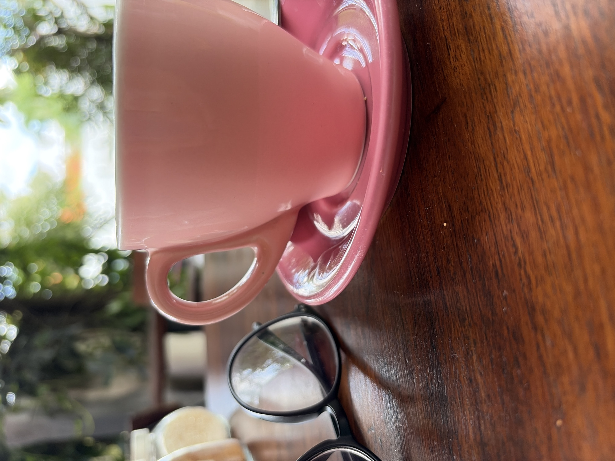 The image shows a close-up of a pink coffee cup and saucer on a wooden table. To the left of the cup are a pair of black-rimmed glasses. In the blurred background, greenery and bokeh lights suggest an outdoor setting, possibly a cafe or restaurant in North Kuta, Indonesia. There are no people visible in the image. The focus is on the coffee cup, saucer, and glasses, with the wooden table surface taking up the lower portion of the frame.
