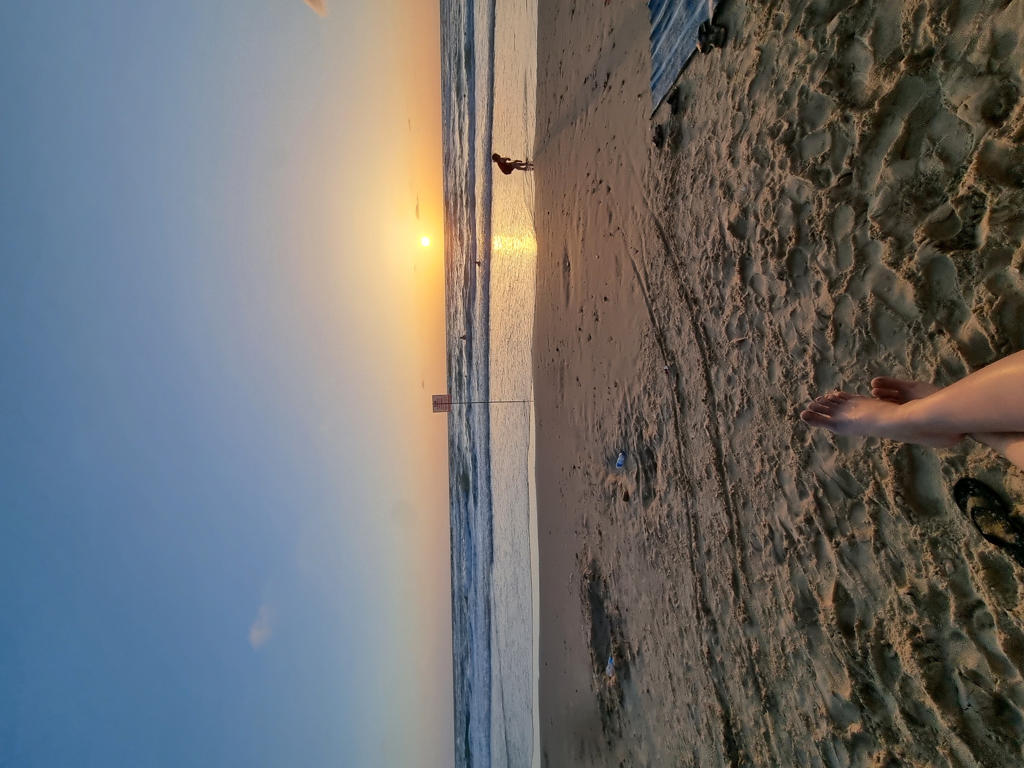 The image depicts a beach scene at sunset, with the horizon running diagonally across the frame. The sky transitions from a clear blue on the left to a warm orange hue near the horizon, where the sun is setting. The ocean waves are visible, with sunlight reflecting off the water surface. A lone person is walking along the shoreline, silhouetted against the sunset. On the right side of the image, the sandy beach shows footprints and textures. A pair of bare feet are partially visible in the bottom right corner. Given the mention of Tel-Aviv, Israel, it is likely this scene takes place on one of the city's Mediterranean beaches.