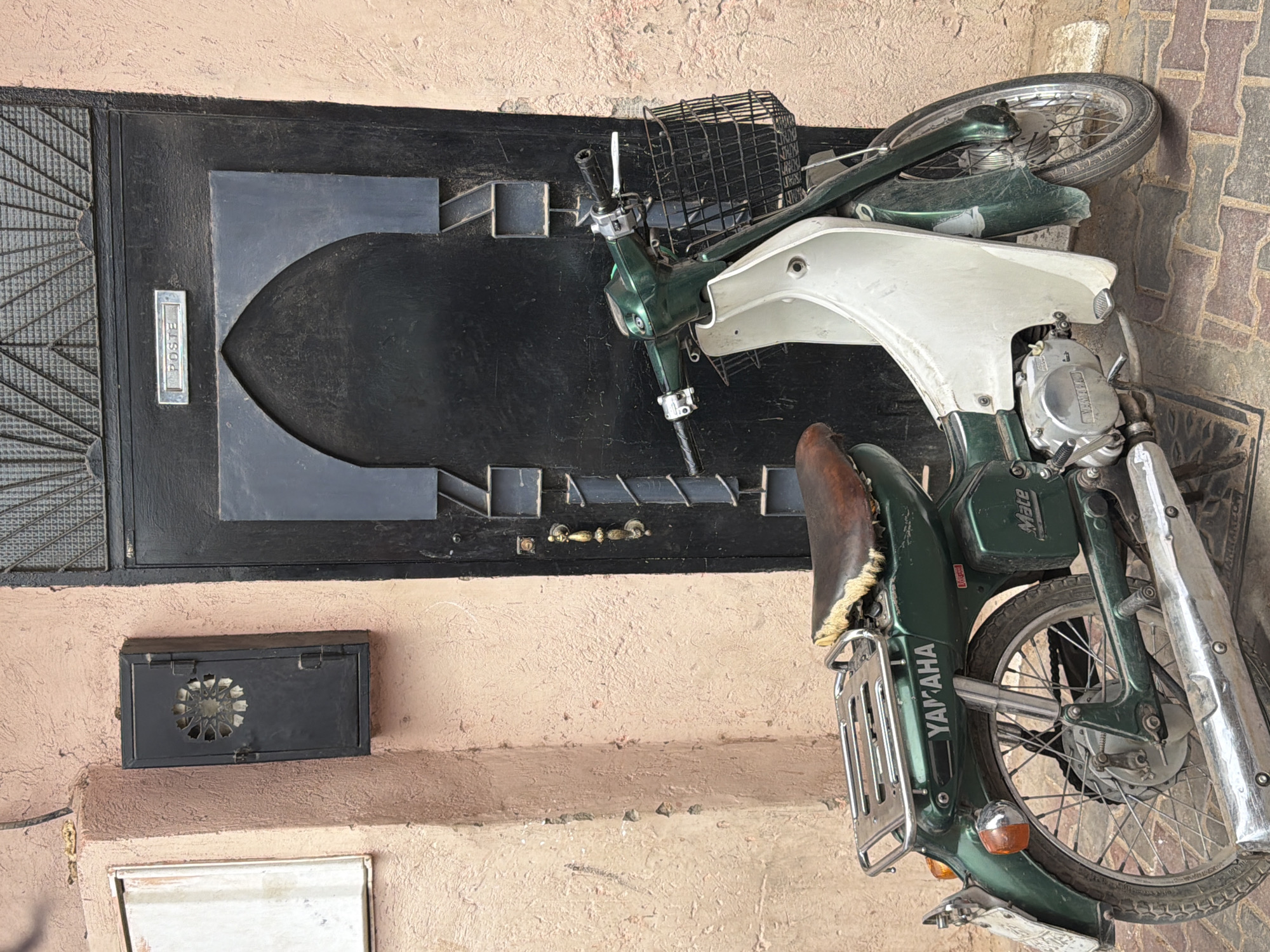 This image captures a close-up view of a vintage green Yamaha Mate moped parked in front of a dark, ornate metal door. The moped is the central subject, leaning slightly to the right. Its features include a black wire basket on the handlebars, a cream-colored front fairing, a worn brown leather seat with a fringed edge, and chrome accents. The "YAMAHA" logo is prominently displayed on the side panel, and the word "Mate" is visible on a smaller panel above the rear wheel.
To the left of the moped, the door exhibits a patterned metal grating and a rectangular silver mailbox labeled "POSTE." Below the door and to the left of the moped's rear wheel is a black rectangular ventilation grate with a decorative, star-like pattern.
The moped and the door are set against a textured, pale pink wall, with a tiled surface visible on the right side of the frame. The lighting suggests it is daytime, with natural light illuminating the scene. The overall impression is one of a quiet, perhaps residential, setting in Marrakesh, Morocco, where everyday objects like the moped and the distinctive doorway contribute to the local atmosphere. There is no visible activity or event taking place; the scene is static, focusing on the details of the moped and its surroundings.