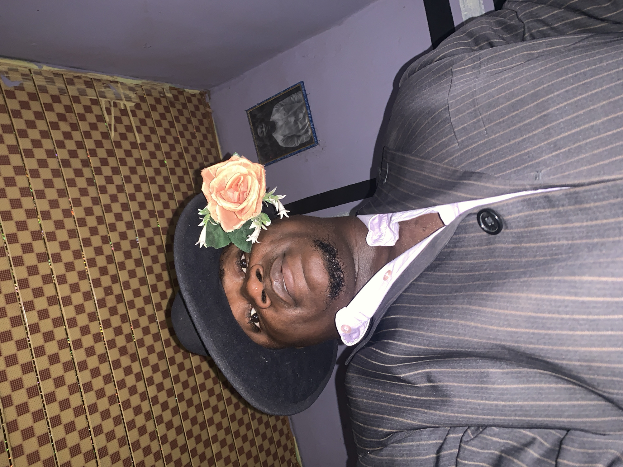 The image shows a man wearing a dark pinstripe suit, a light purple shirt, and a black hat with a peach-colored rose and white flowers attached. He is indoors, standing in a room with a brown and tan patterned wall covering. On the lilac-painted wall behind him is a framed black and white photograph. The exact location in Akwanga, Nigeria, is not discernible from the image.