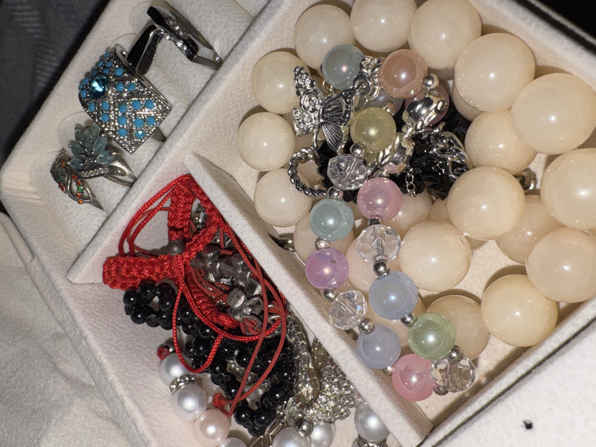 The image shows a jewelry box filled with various accessories. In the top right compartment, there are several rings, one of which is silver with a large turquoise stone and smaller blue stones. Next to it is a silver ring with a floral design in blue and green stones. Another silver ring is visible with orange and green stones. In the left compartment, there are strands of pearls, black beads, and a red braided cord. The bottom compartment contains several bracelets made of large, smooth, light-colored beads, some with pastel-colored beads and clear crystal beads interspersed, as well as charms. The jewelry is neatly organized within the compartments of the jewelry box.
