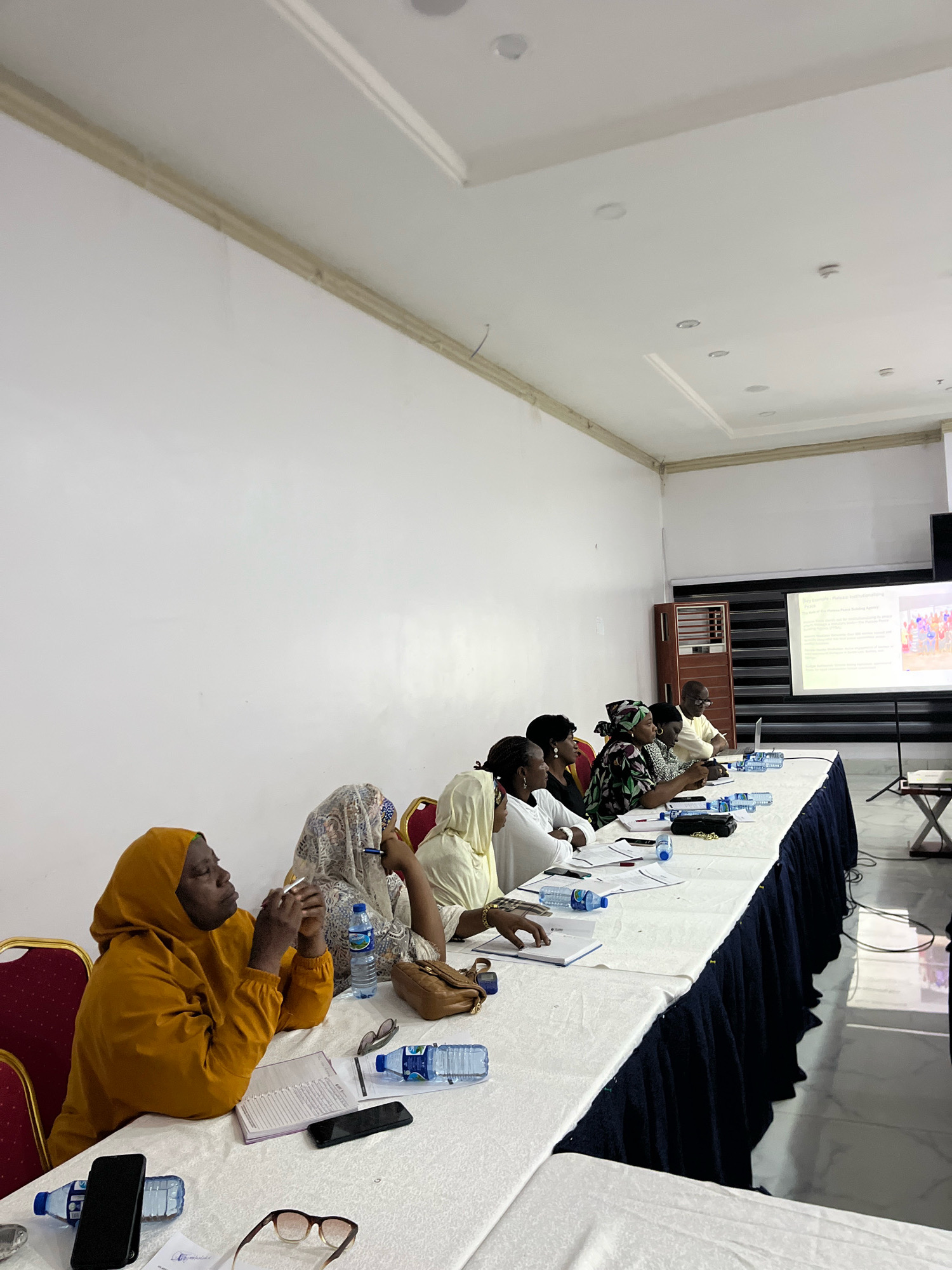 The image depicts a meeting in progress, likely at a conference center or a training facility in Jos, Nigeria, given the presence of a projector screen displaying text and an image. A group of individuals, predominantly women, are seated at a long, white-draped table. Some are attentively looking towards the front, possibly at the projector screen, while others are engaged in note-taking or listening.
In the foreground, several water bottles, notebooks, pens, and mobile phones are scattered on the table, indicating a session of learning or discussion. A woman in a bright yellow hijab is seated on the left, looking upwards, with a notebook and pen in front of her. To her right, another woman in a patterned headscarf is also seated, with a pen in her hand. Further down the table, a man is visible, appearing to be a participant or facilitator.
The room features white walls and a drop ceiling with recessed lighting. A projector screen, mounted on a stand, is visible on the right side of the image, displaying text that suggests a presentation title like "Jos Example: Plateau SDG/Monitoring Project" and bullet points outlining recommendations or findings. The overall atmosphere suggests a formal gathering or workshop. The lighting in the room is bright, with some shadows cast by the individuals and objects on the table.