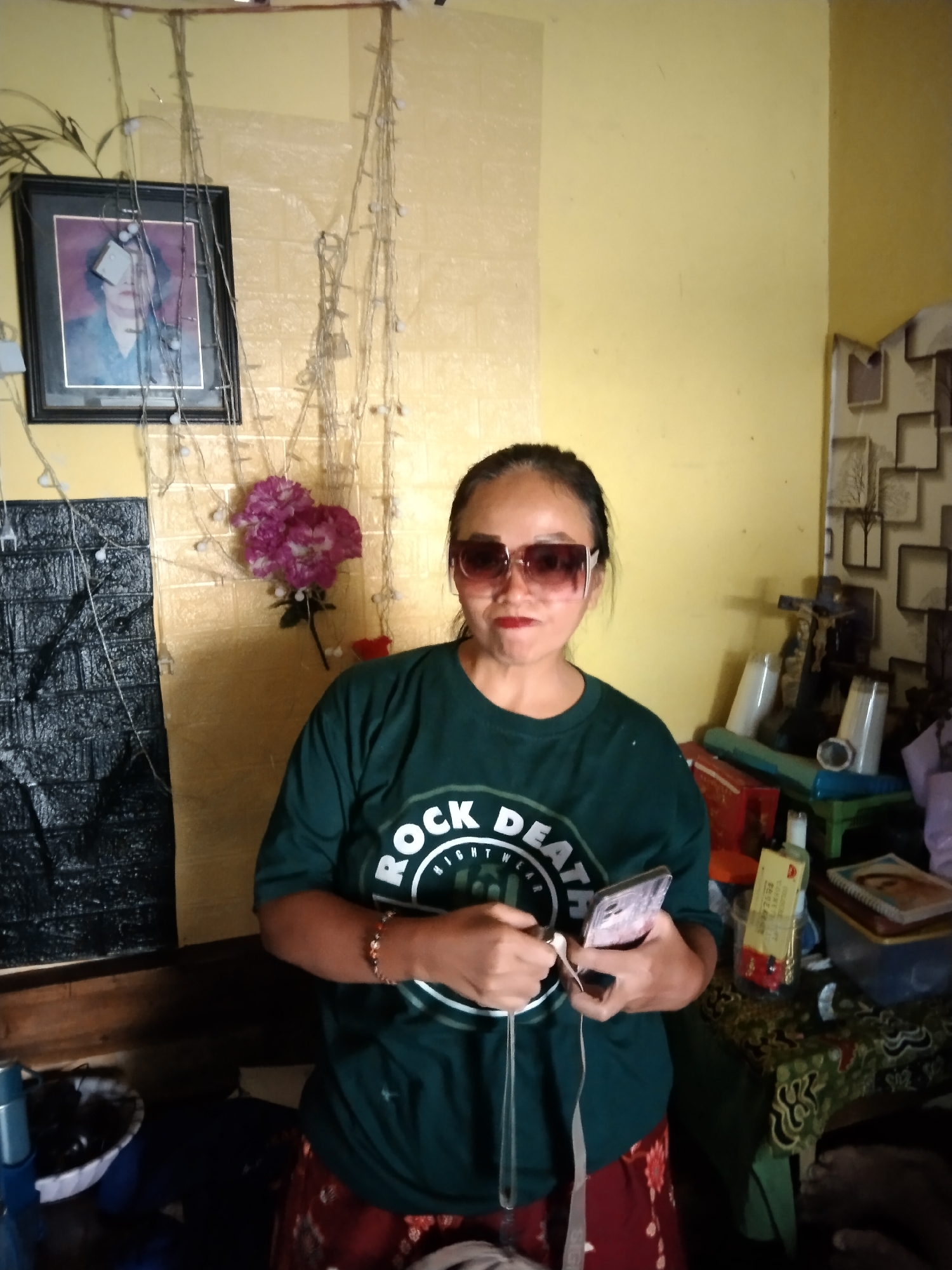A medium shot captures an Indonesian woman in Magelang, Indonesia, standing in a room with yellow walls. She is wearing large, square sunglasses, a dark green t-shirt with a "ROCK DEATH" logo, and a patterned red skirt. The woman is holding a smartphone and earbuds in her hands. Behind her, the wall is decorated with strings of small lights and a framed portrait. To her right, a table is laden with various items, including cups, bottles, and books. The overall impression is of a casual indoor setting.