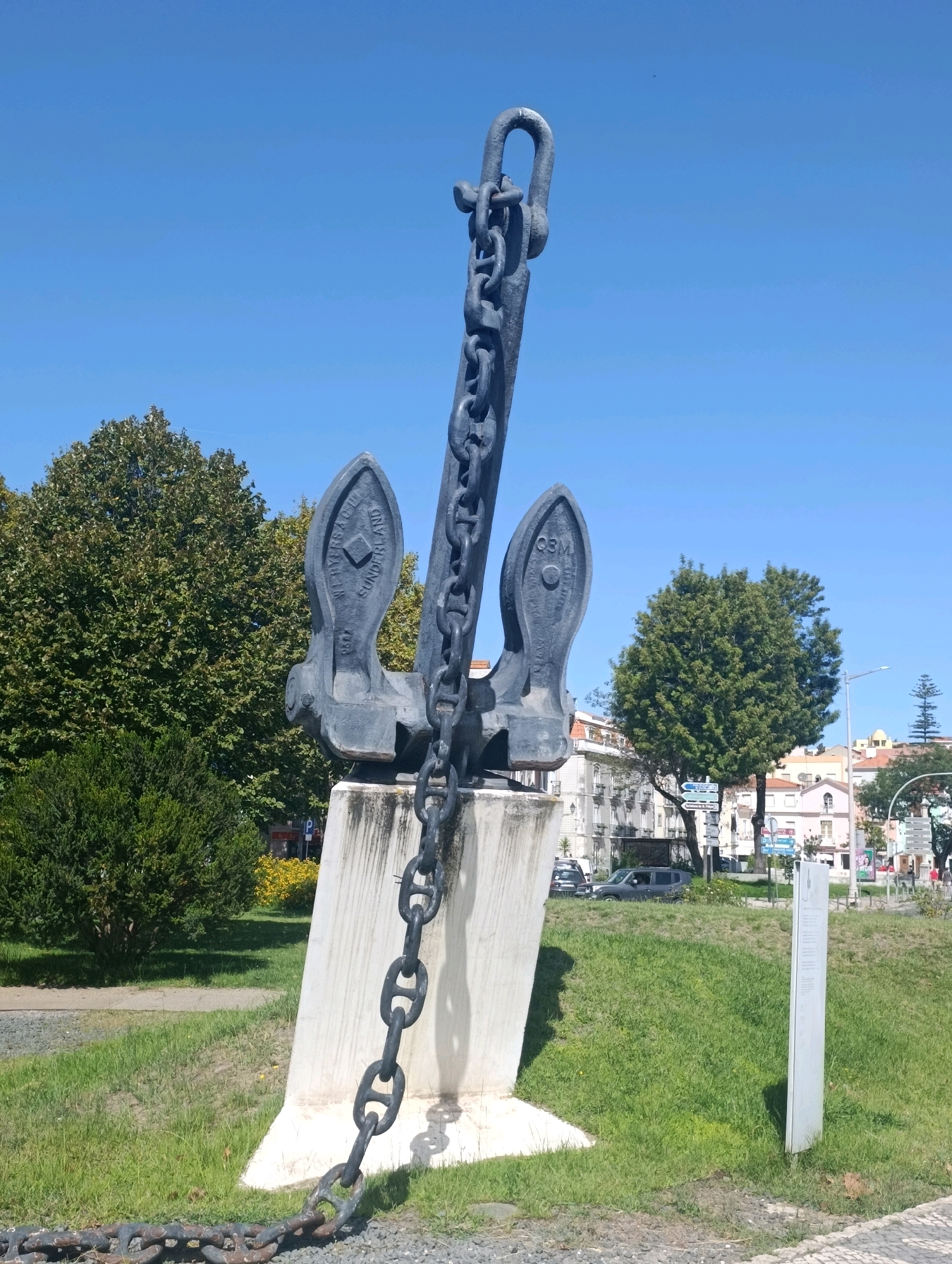 The image shows a large, dark gray anchor sculpture mounted on a concrete base in a park-like setting. The anchor is made of metal and features a long, thick chain attached to its top. The chain drapes down and extends across the ground in front of the sculpture. The base of the anchor has an inscription that reads "WE MYERS & Co SUNDERLAND 1907" on one side and "Q3M" on the other. Behind the sculpture, there are trees, grass, and buildings in the background. The sky is clear and blue. This monument is located at the roundabout of Avenida do Engenheiro Edgar Cardoso in the city of Setúbal, Portugal. The scene depicts a static monument in an urban green space on a sunny day.