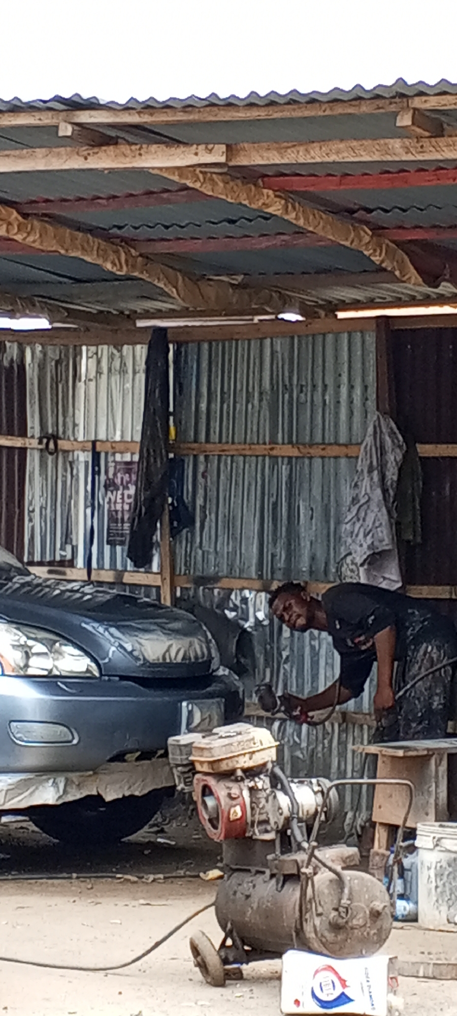 The image depicts an active, semi-enclosed auto body workshop, likely situated in Lagos, Nigeria, given the rustic construction and type of activity. A man, dressed in dark, paint-splattered clothing, is focused on spray painting the front bumper of a dark-colored SUV, resembling a Lexus RX model. He holds a spray gun and leans over the vehicle, which has its hood and other sections covered with masking material to protect unpainted areas.
The workshop structure is utilitarian, with walls and a roof constructed from corrugated metal sheets supported by rough wooden beams and planks. Some parts of the roof show signs of patching. On the dirt floor in the foreground, a robust, aged air compressor is positioned. It features a visible red engine block on top, a cylindrical tank below, and small wheels for mobility. Hoses connect it to the spray gun.
In the background, hanging on the corrugated metal wall, are dark garments or rags. A partially obscured poster or sign on the wall shows the text fragments "WED" and "NEC". Near the compressor, a white bag or container is visible on the ground, clearly displaying the brand name "IDEAL" in blue lettering, accompanied by a red and blue logo. The scene conveys a sense of industrious work in a practical, informal setting during daytime.