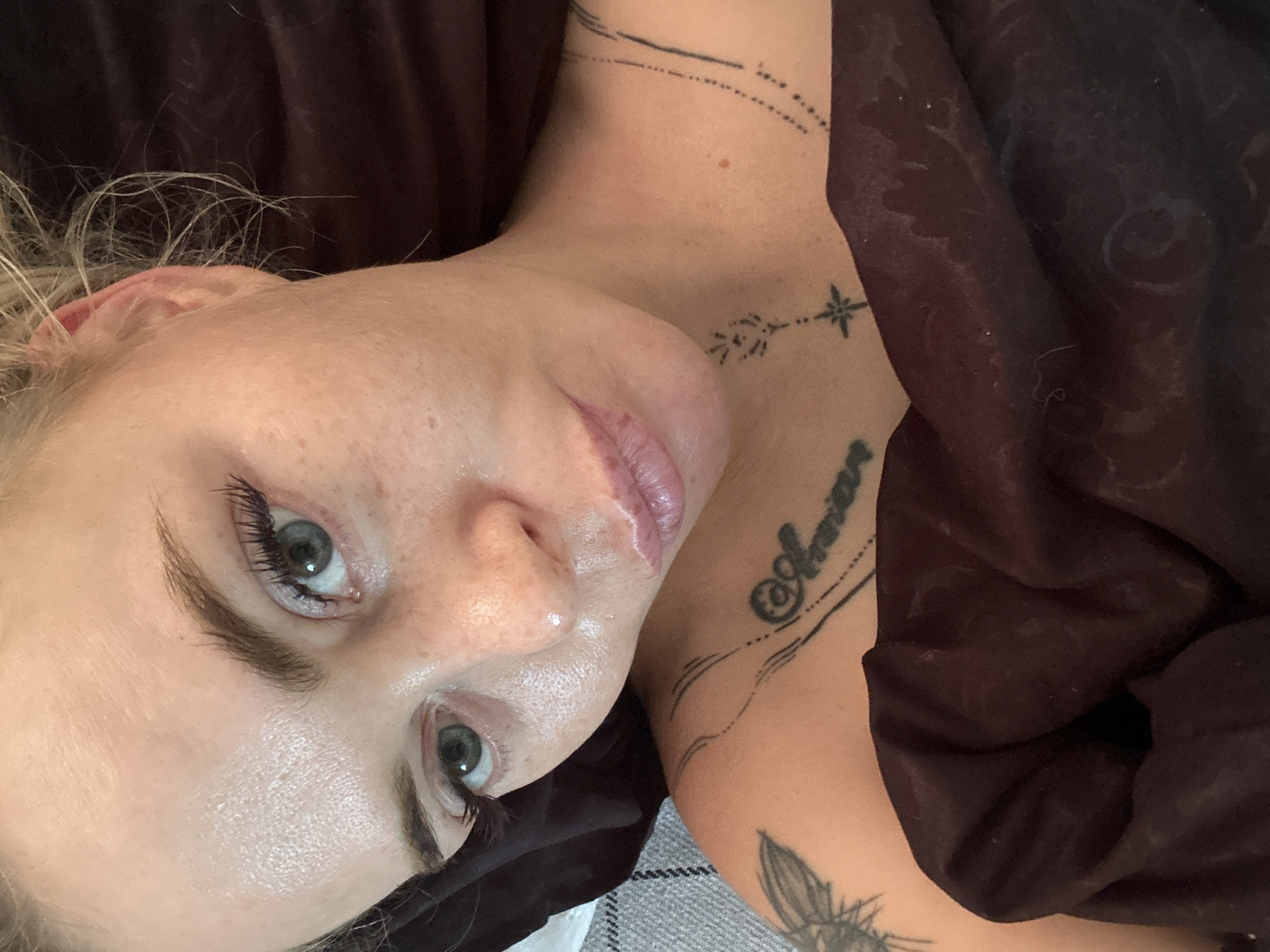 An overhead, close-up, selfie-style shot shows a light-skinned person lying in bed. The person has fair skin, freckles, and blonde hair that is slightly messy. Their eyes are a light blue-green, and they are wearing mascara. Their lips are slightly parted and appear to have lip gloss or balm on them. The person is wearing a dark-colored blanket, which is covering their chest. There are two visible tattoos on the person's shoulder and upper chest. One tattoo on the shoulder is a detailed wing, and another on the chest is the word "Amor" written in script. The background is dark and out of focus, with a glimpse of a grey and white checkered surface on the left.