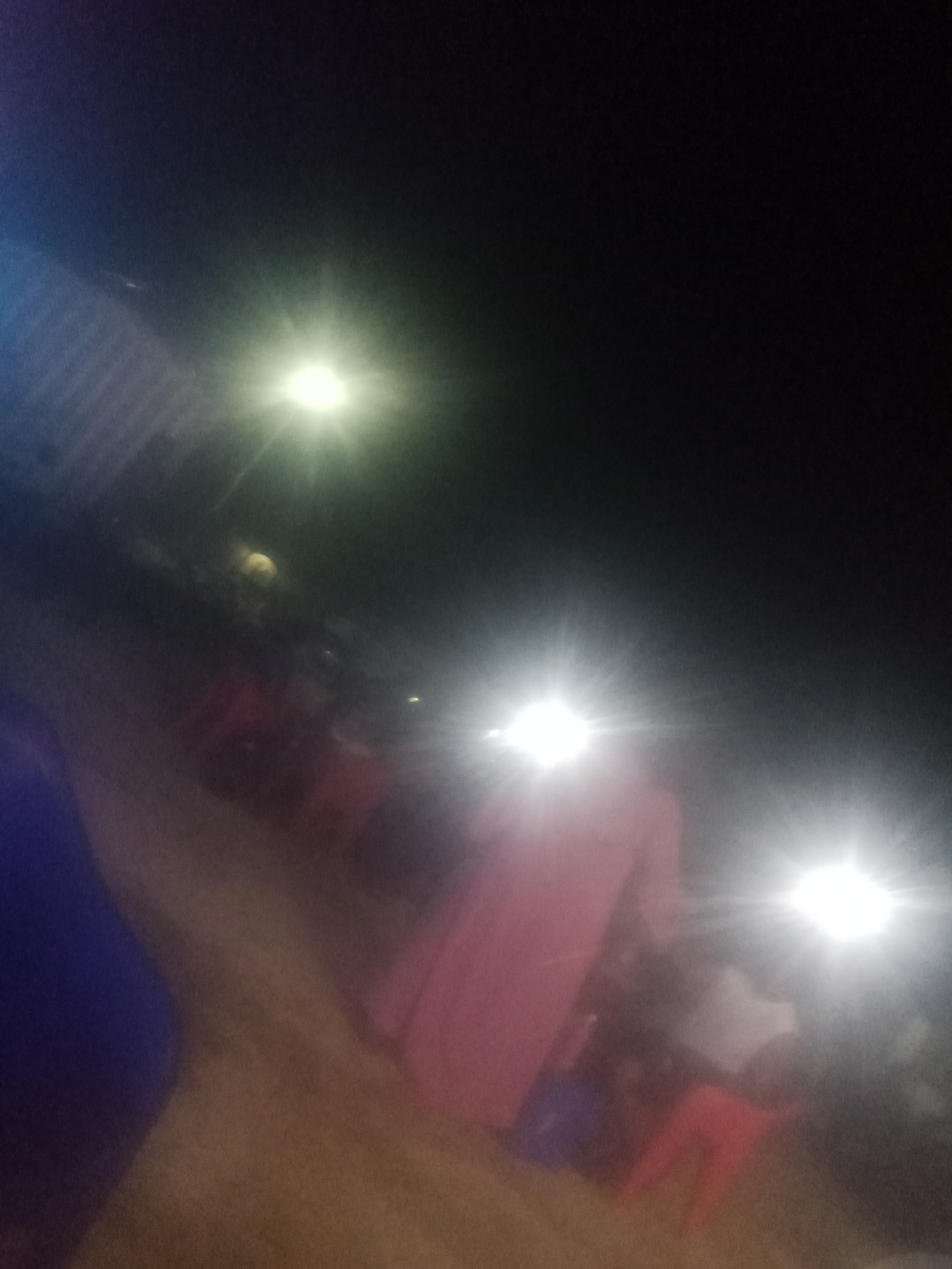 The image depicts a nighttime outdoor gathering, likely in Jalingo, Nigeria, characterized by multiple artificial light sources causing significant glare. In the foreground, a person in a long, pink garment is partially visible, appearing to be standing. Behind them, other individuals are present, though their features are obscured by the low light conditions and glare. The environment appears to be an open area, possibly a street or courtyard, with a sandy or dirt ground. The background is dominated by darkness, punctuated by the bright lights. A striped structure, possibly a building wall or fence, is visible on the left side, illuminated by a bright light source. The overall impression is one of a community event or social gathering taking place after dark, with limited visibility due to the lighting.