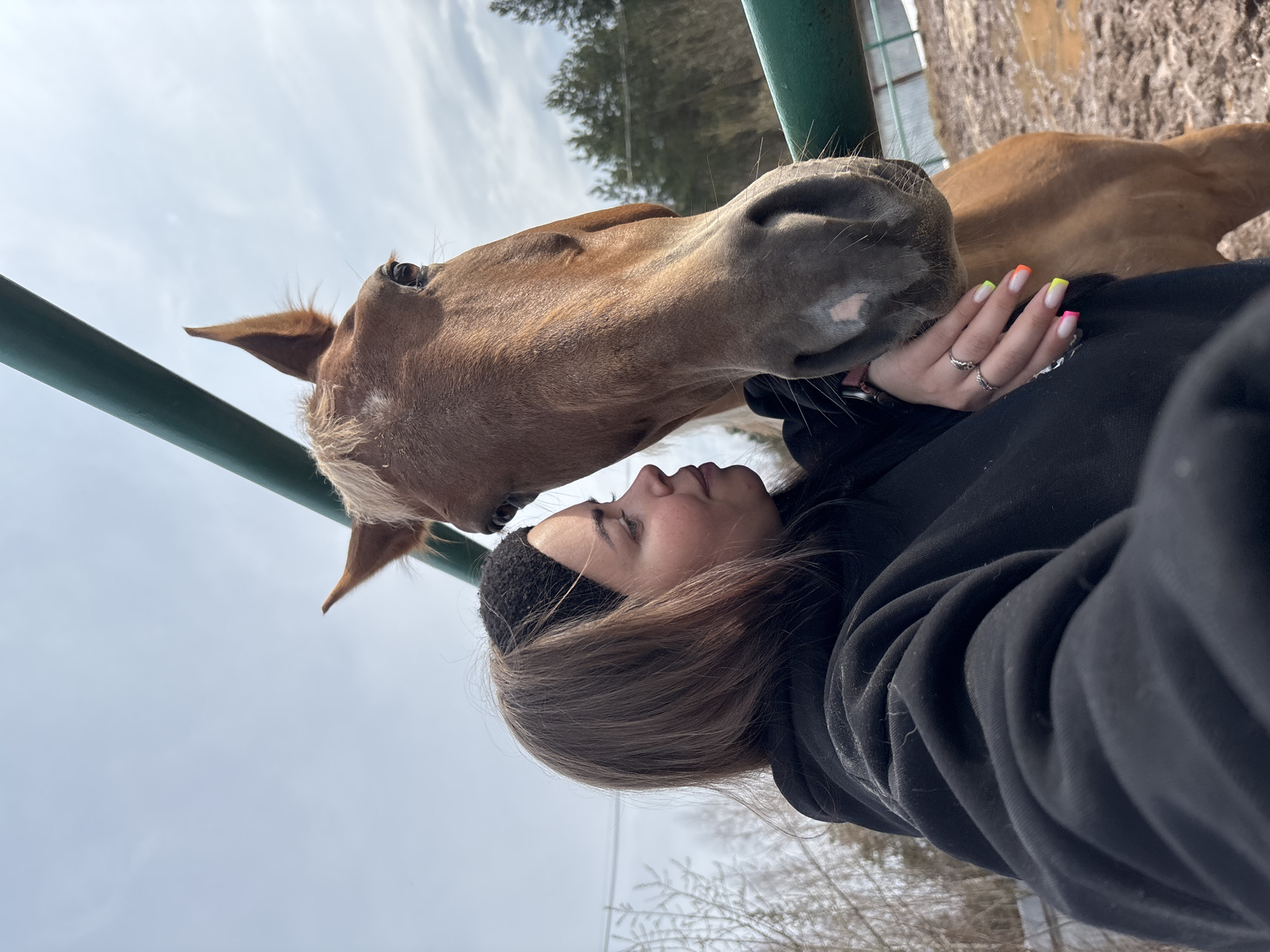 A woman in a black hooded sweatshirt and a black headband is interacting affectionately with a light brown horse near what appears to be a green metal fence. The woman's face is close to the horse's head, suggesting a gentle connection. The horse's muzzle is touching the woman's hand, which has painted nails with a light base and colored tips. A portion of the horse's body extends behind the woman. The setting appears to be outdoors, with trees visible in the background under a cloudy sky. The foreground shows evidence of winter conditions, with possible traces of snow or frost. There is no specific information to determine that the location is Русский Вожой, Russia.