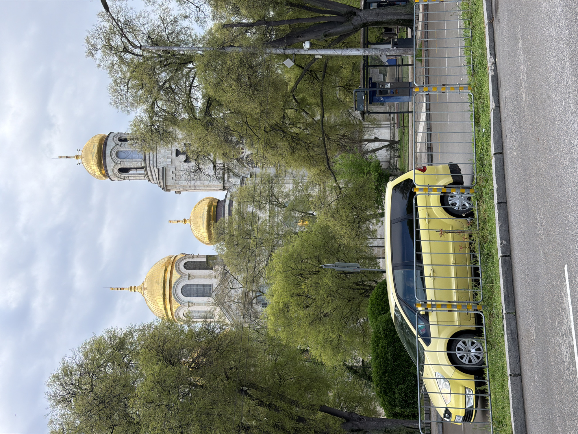 This daytime image captures a grand Eastern Orthodox cathedral or church, partially framed by vibrant green trees, with a yellow car parked in the foreground.
The central subject is a large religious building, characterized by its ornate white stone architecture and multiple golden onion domes. Two prominent domes are visible: a taller, more slender one atop a bell tower in the background, and a broader, slightly lower one in the mid-ground. Both domes feature arched windows and architectural detailing, gleaming under a bright, overcast sky.
Numerous deciduous trees with fresh, light green leaves, indicative of springtime, are prominent throughout the scene. They partially obscure the church structure and extend into the foreground, creating a natural frame.
In the right foreground, a light yellow minivan or station wagon is parked on a grassy strip. It is separated from a grey asphalt road by a temporary metal barrier fence. This fence is made of grey horizontal and vertical bars, with some sections highlighted by yellow and black striped tape, suggesting a safety or event boundary. The car's front end and side are visible, including its wheels and headlights. The road itself has a single white painted line and is bordered by a concrete curb.
The scene appears quiet, suggesting a calm day with diffused light from a cloudy or partly cloudy sky. The architectural style of the church strongly points to an Eastern Orthodox context, aligning with the provided location of Varna, Bulgaria. No people or discernible text are visible in the image.