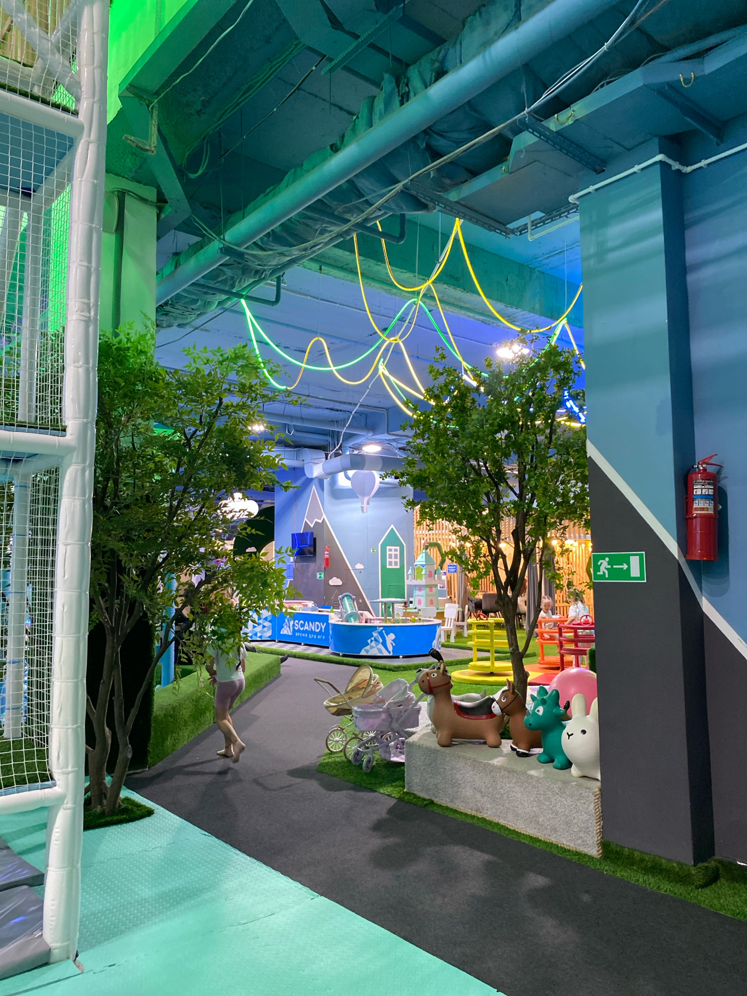 The image depicts the interior of the "SCANDY" children's play center in Izhevsk, Russia. The scene is brightly lit with a combination of artificial lights and neon accents. In the foreground, a white safety net structure is visible on the left, typical of indoor playgrounds. Artificial trees with green foliage are strategically placed throughout the space.
A person, a young woman, is walking away from the camera on a dark grey path, wearing a light-colored t-shirt and pink shorts. Her legs and feet are visible as she walks barefoot. To her right, a vintage-style baby stroller with a light-colored hood is parked.
Further into the play center, several colorful children's play structures are visible. On a grey stone-like ledge, there are four inflatable toys: two brown horses, a green dragon, and a white rabbit. Beyond these toys, a carousel with red and yellow seats is set up, with children playing on it. In the background, the walls are decorated with murals depicting mountains and clouds. A red fire extinguisher is mounted on a blue wall on the right side of the image, with a green emergency exit sign below it. The ceiling is high and industrial, with exposed pipes and conduits, and decorated with wavy yellow and green neon lights. The floor is a mix of dark grey pathways and light green, textured play mats.
