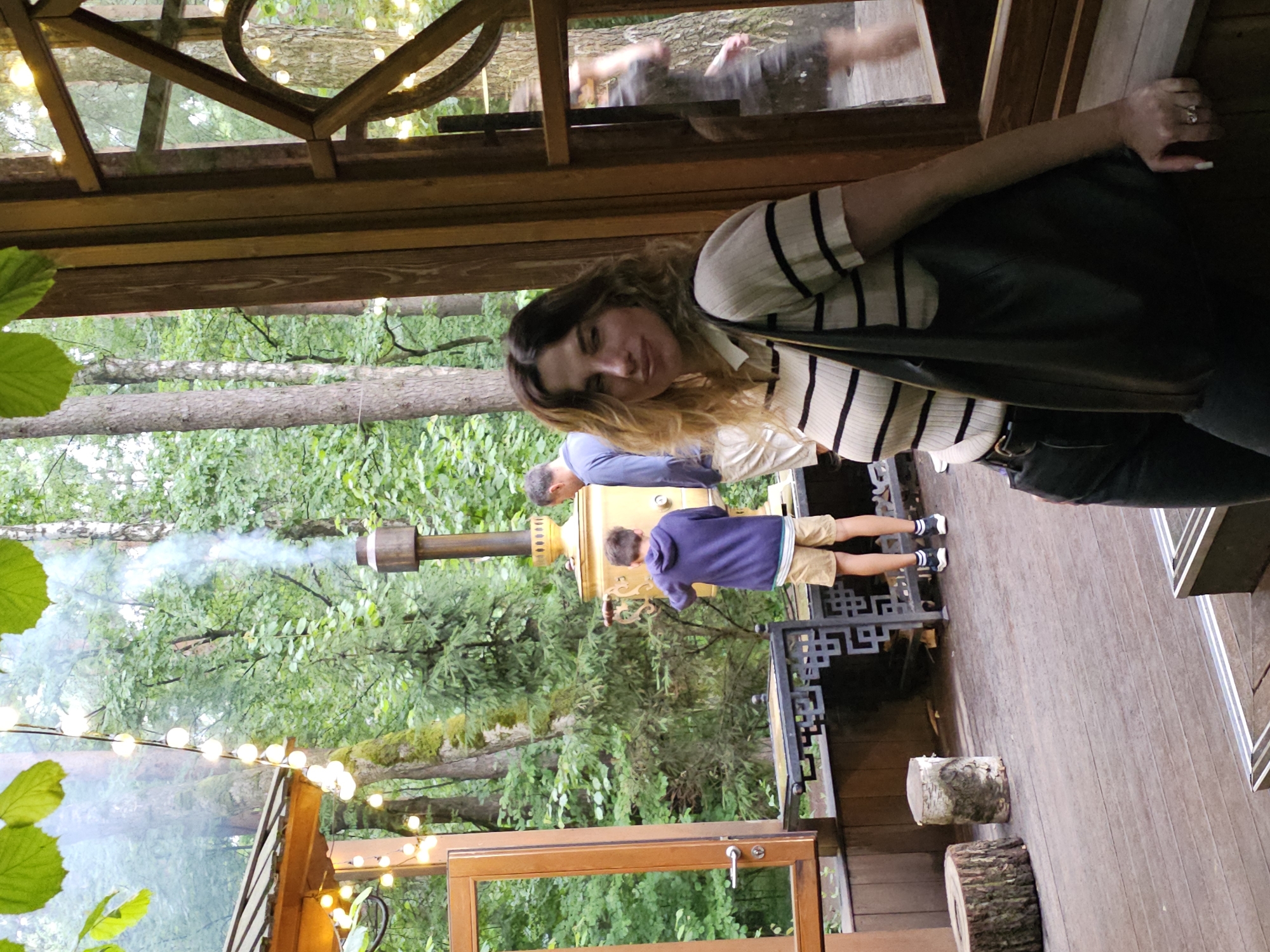 In Nemchinovo, Russia, a young woman with wavy brown hair, wearing a striped shirt and black pants, stands near a wooden structure. She carries a dark bag over her shoulder. In the background, a boy in a purple hoodie and shorts stands by a large, golden samovar, from which smoke rises. A man in a blue shirt and beige shorts is beside the boy, observing the samovar. The scene is set in a wooded area, with trees and foliage surrounding the structure, illuminated by strings of lights. Two tree stumps are visible on the wooden deck. A blurred figure is seen through a window on the right side of the image.