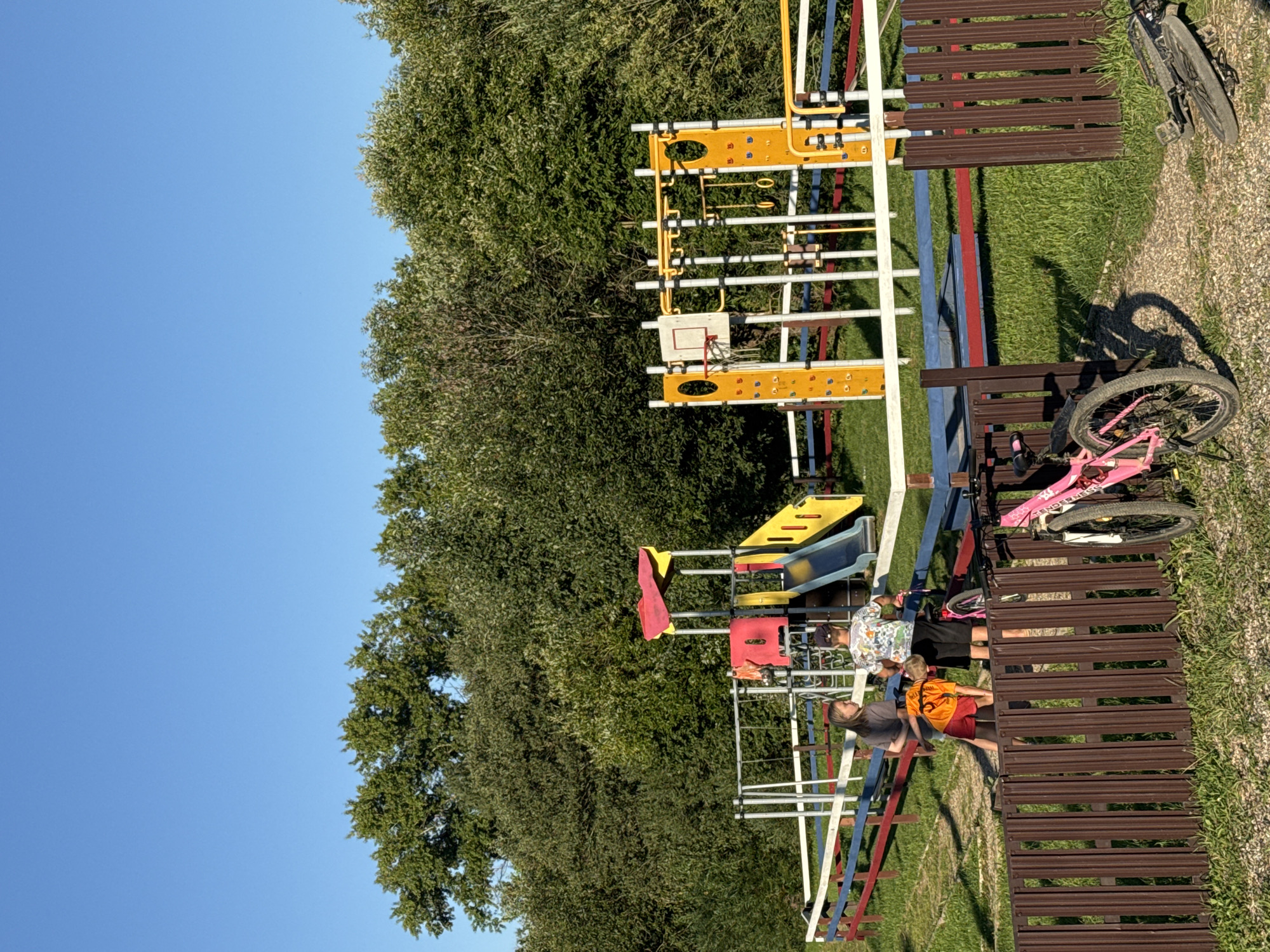 The image shows a sunny day at an outdoor playground with lush green trees in the background. In the foreground, a brown wooden fence runs horizontally across the bottom of the frame, with two bicycles parked nearby. One is a pink bicycle, upright and partially in view on the left, while the other is a darker bicycle lying on its side on the right.
Beyond the fence, a colorful playground structure features red and yellow slides, swings, and climbing equipment. Several children are interacting near the playground. Two children are standing on the left side, while a third child in an orange shirt is bending down in the middle. The playground is bordered by a fence with horizontal red, white, and blue planks.
The scene is set in an outdoor park, likely a recreational area in Krasnaya Presnya, Russia, judging by the context. The sky above is a clear, bright blue.