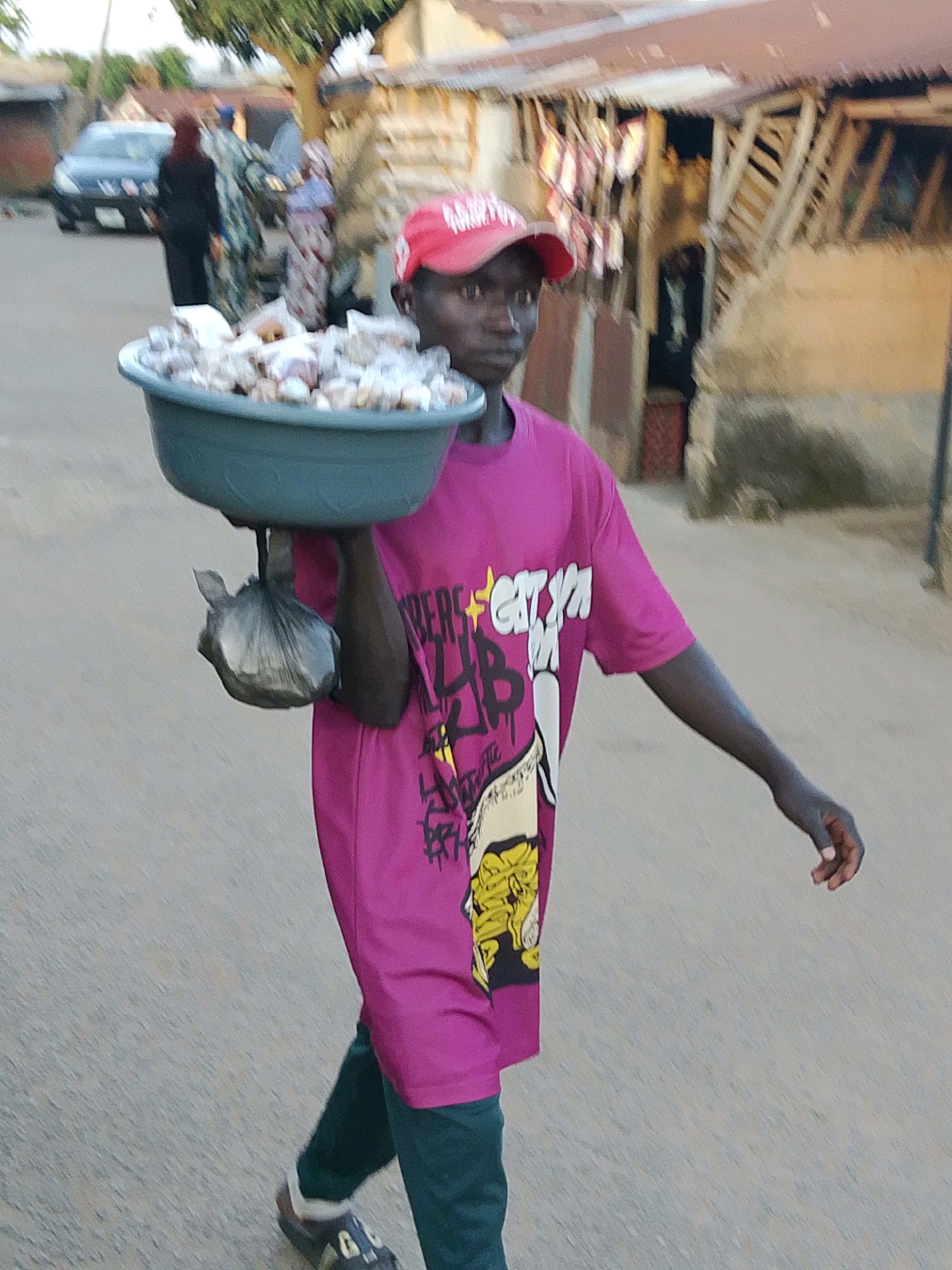 The image depicts a young person wearing a bright pink t-shirt and dark green trousers, carrying a large grey basin filled with goods on their head. The person has a red baseball cap on and is walking down a paved road. In the background, there are buildings with corrugated iron roofs and some vegetation. To the left, a dark-colored car is visible. Other people, including one in a black headscarf and another in a blue headscarf, are also present in the background, walking away from the viewer. The overall scene suggests a busy street or market area in a city. The style of the buildings and the general atmosphere are consistent with an urban environment in Nigeria, specifically the city of Jos. The goods in the basin appear to be individually wrapped items, possibly food or small wares.