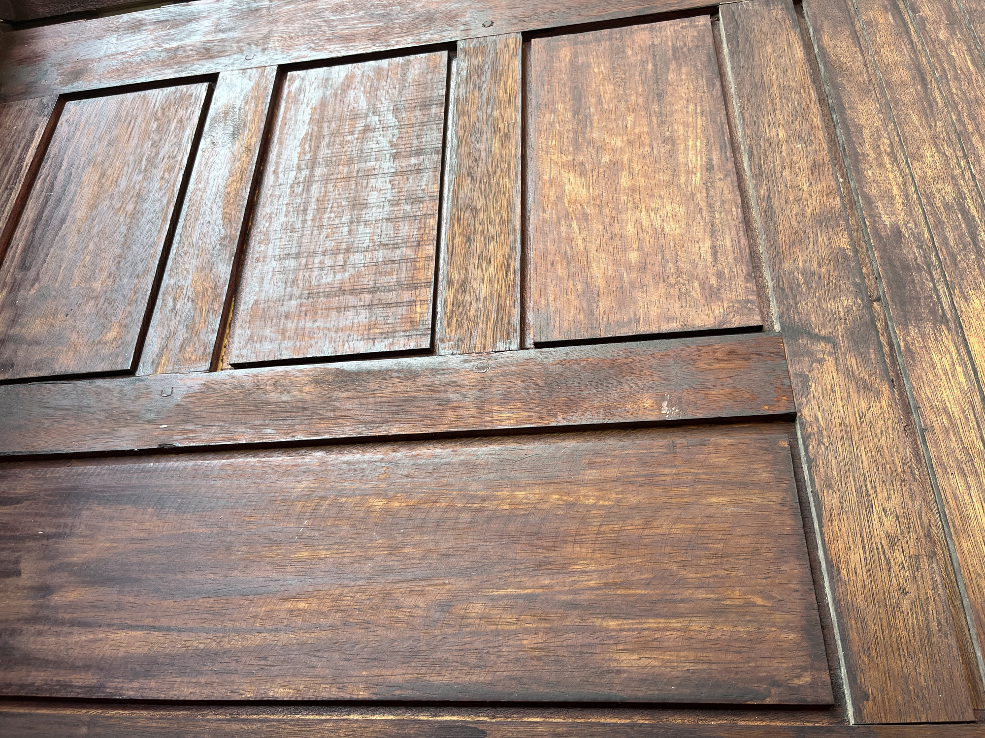 This close-up, angled photograph depicts a section of a dark reddish-brown wooden structure, likely a door or wall panel, in Lhokseumawe, Indonesia. The surface is composed of multiple wooden elements, displaying prominent grain patterns and variations in color. On the left, a large, vertically oriented panel exhibits visible wood grain running predominantly lengthwise. To the right, a narrower vertical wooden stile separates this panel from a section featuring three distinct horizontal wooden slats or louvers, each showcasing horizontal grain patterns and color differences, including areas of lighter reddish-brown and darker, almost black, streaks. The wood appears weathered, with a moderately textured surface and some areas displaying a slight sheen. No individuals or activities are present within the frame.