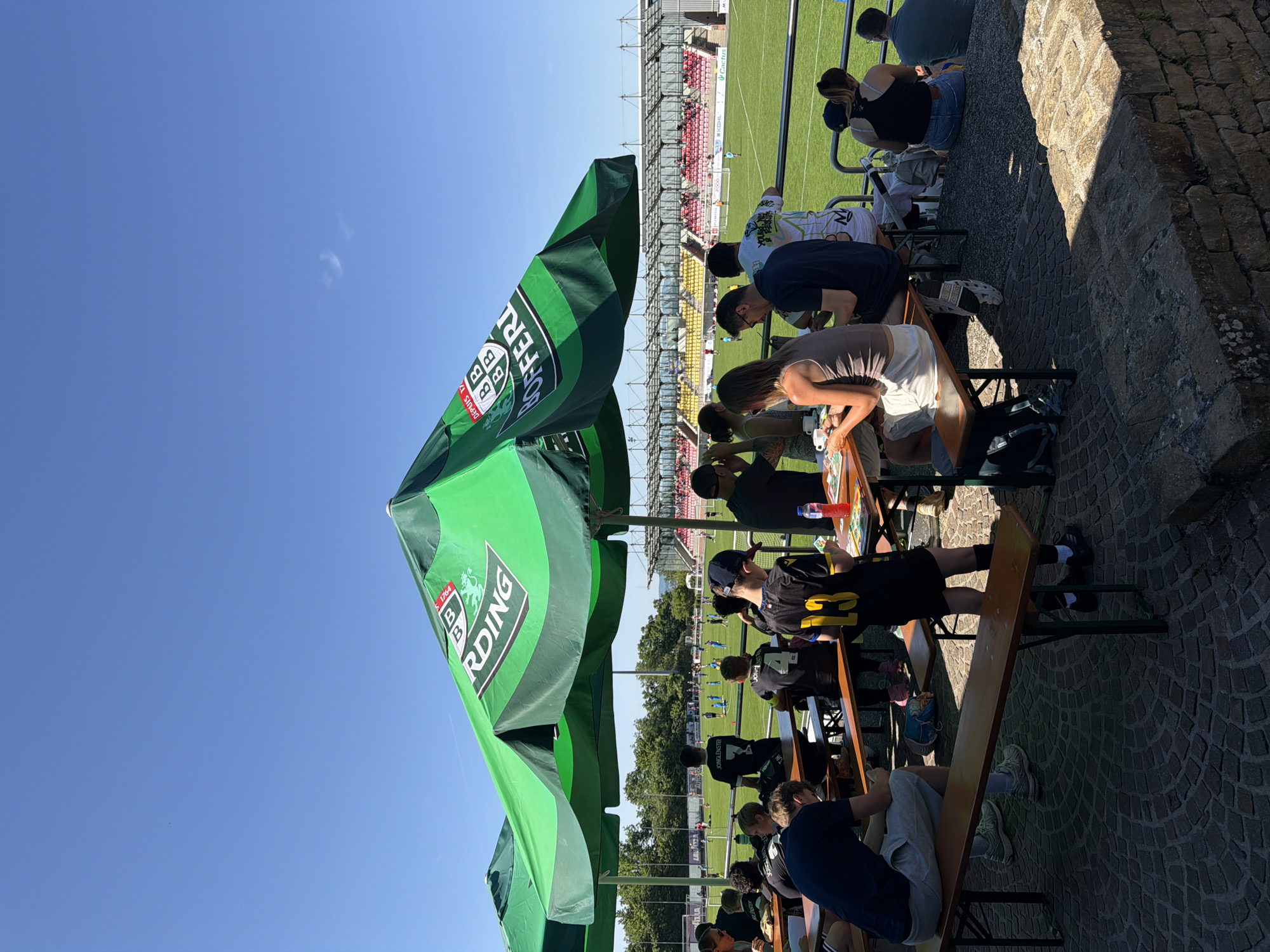 The image shows a group of people sitting at tables under green umbrellas. There are two rows of tables, with people sitting on benches facing the soccer field. On the field, a soccer game is taking place. In the background, a stadium with red and yellow seats is visible. This scene is likely in Fentange, Luxembourg, as indicated in the prompt.