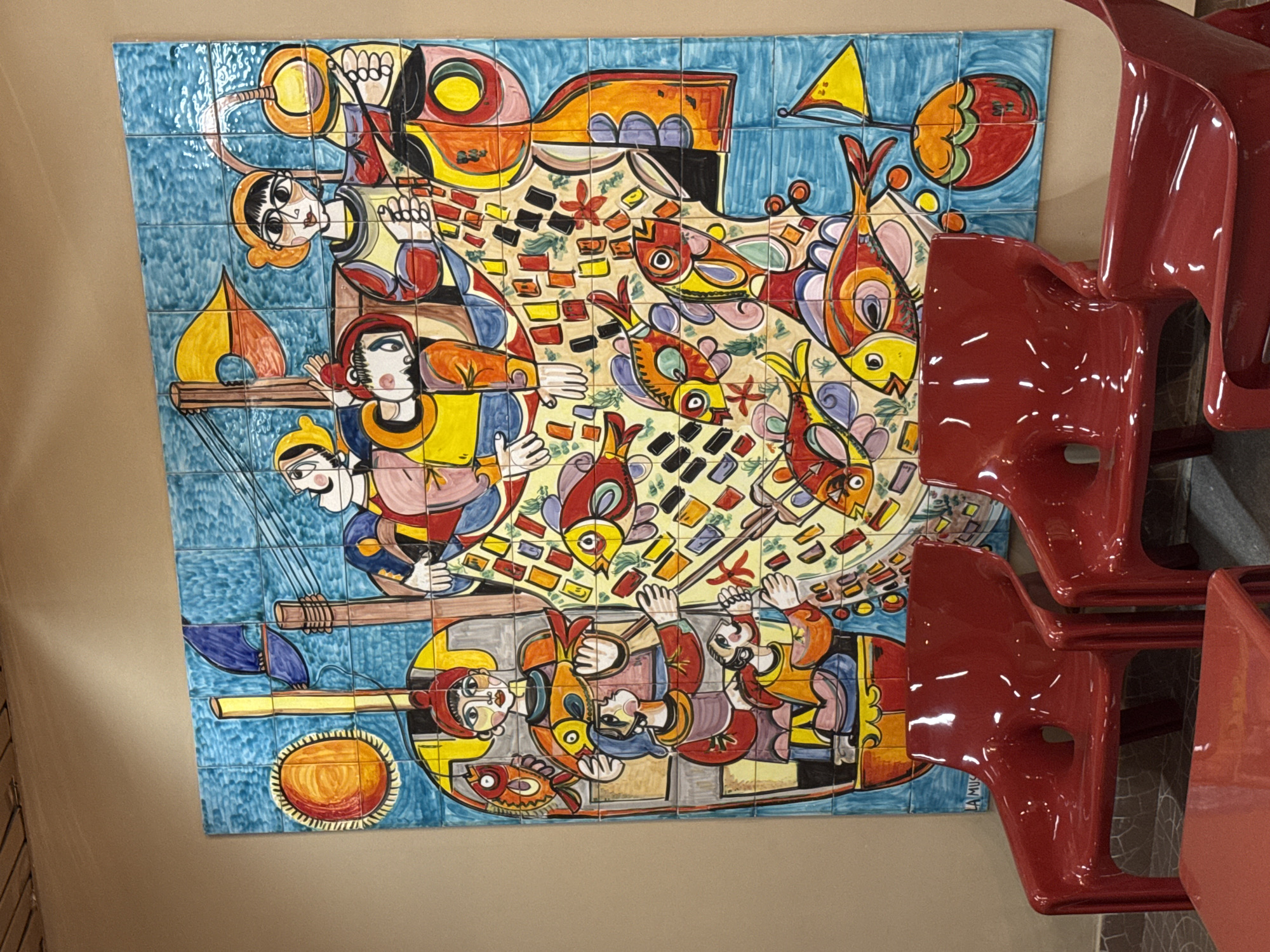A large, colorful ceramic tile mural depicting a fishing scene with multiple figures and fish is displayed on a light brown wall in Isola delle Femmine, Italy. The mural is vibrant and features a cubist-inspired style with bold outlines and geometric shapes. Several red plastic chairs are arranged in front of the mural, some occupied by people, suggesting a public space or a waiting area. The floor is tiled with a dark pattern. The overall impression is a lively and artistic display within a Mediterranean setting.