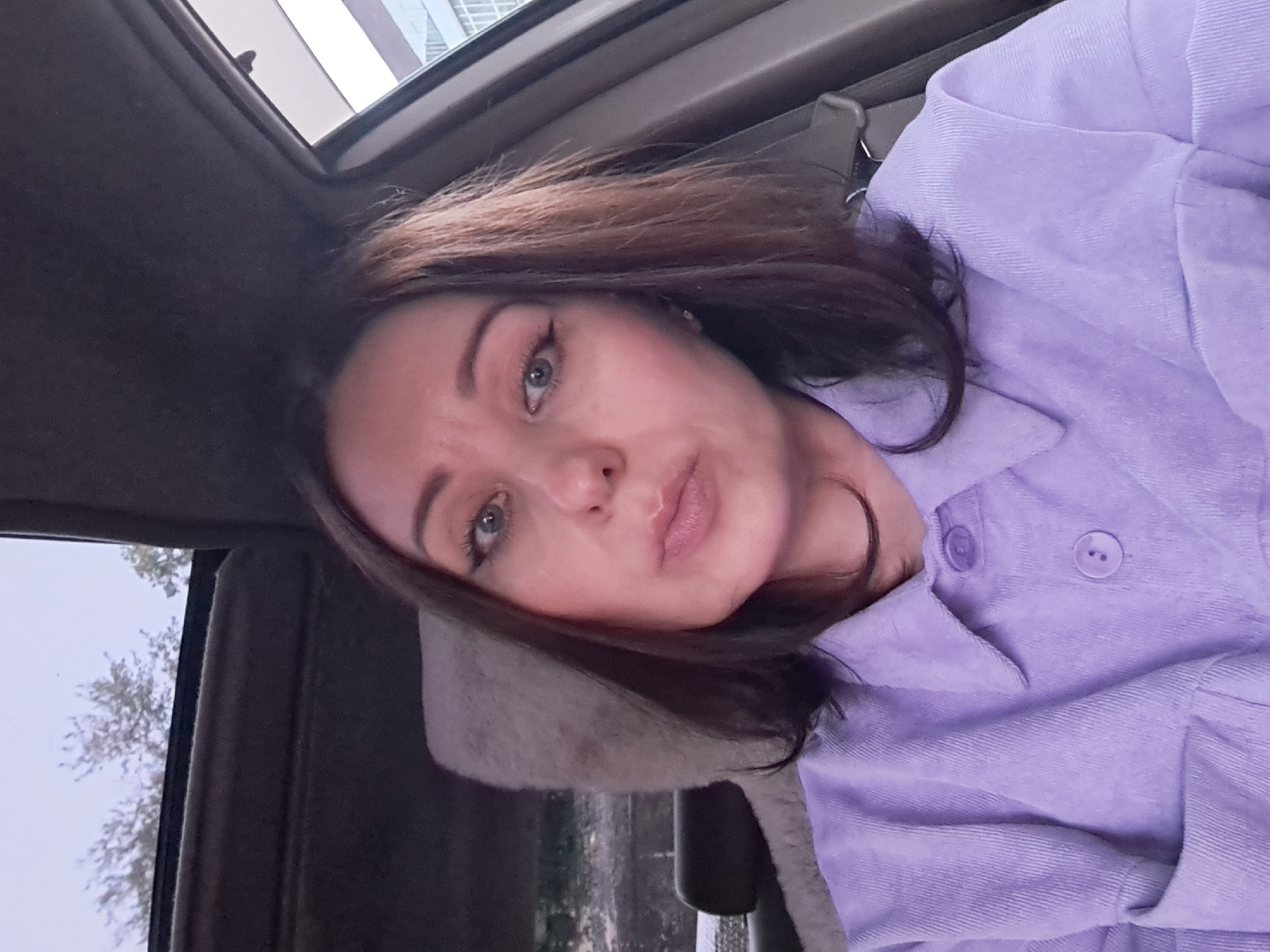 A woman is taking a selfie inside a car, possibly in Irkutsk, Russia. She is wearing a light purple corduroy shirt. The car's interior is visible, including the headrest of the passenger seat and part of the sunroof. The woman has dark brown hair and is wearing makeup. The lighting suggests it might be daytime or dusk. The background outside the car is blurred, showing trees and a building, which could be in a city setting.