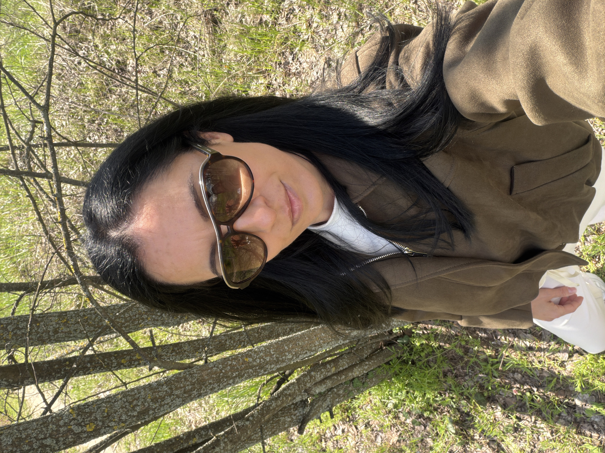 A woman with long, dark hair is seen from a high angle, wearing sunglasses and a brown jacket over a white top. She is outdoors amidst sparse vegetation, with thin tree trunks visible in the foreground. The ground is covered with patches of grass and dry leaves. The location appears to be a natural setting. There are no immediately identifiable landmarks to place the exact location within Pleven, Bulgaria.