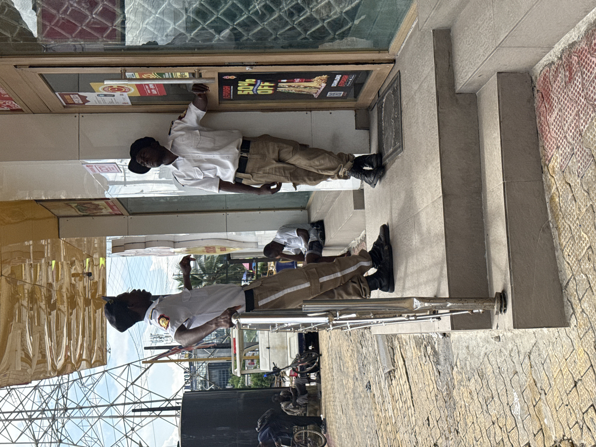 In Itirin, Nigeria, two uniformed security guards stand outside a commercial establishment. The guard on the left, wearing a white shirt and tan pants with reflective stripes, leans against a metal railing and converses with the guard on the right. The second guard, similarly attired, stands by a glass-paneled door with a "50% OFF" advertisement. Behind them, a few individuals are visible in the background; one man sits on steps, appearing tired, while others are further back, near a parked vehicle and a bicycle. The environment suggests a bustling urban setting with visible power lines and a complex infrastructure overhead.