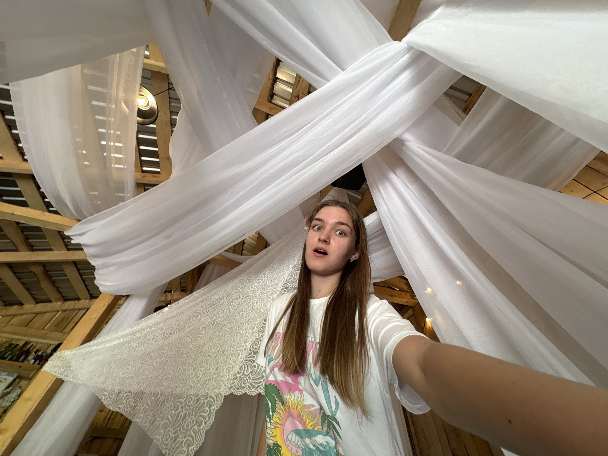 A person with long brown hair is holding a camera at arm's length, taking a selfie from a low-angle perspective. The person is wearing a white t-shirt with a colorful graphic. The background consists of wooden beams and white fabric draped across the ceiling. A light fixture is visible among the beams and fabric. The photograph was likely taken indoors in Sibir', Russia, but there is no clear indication of the city.