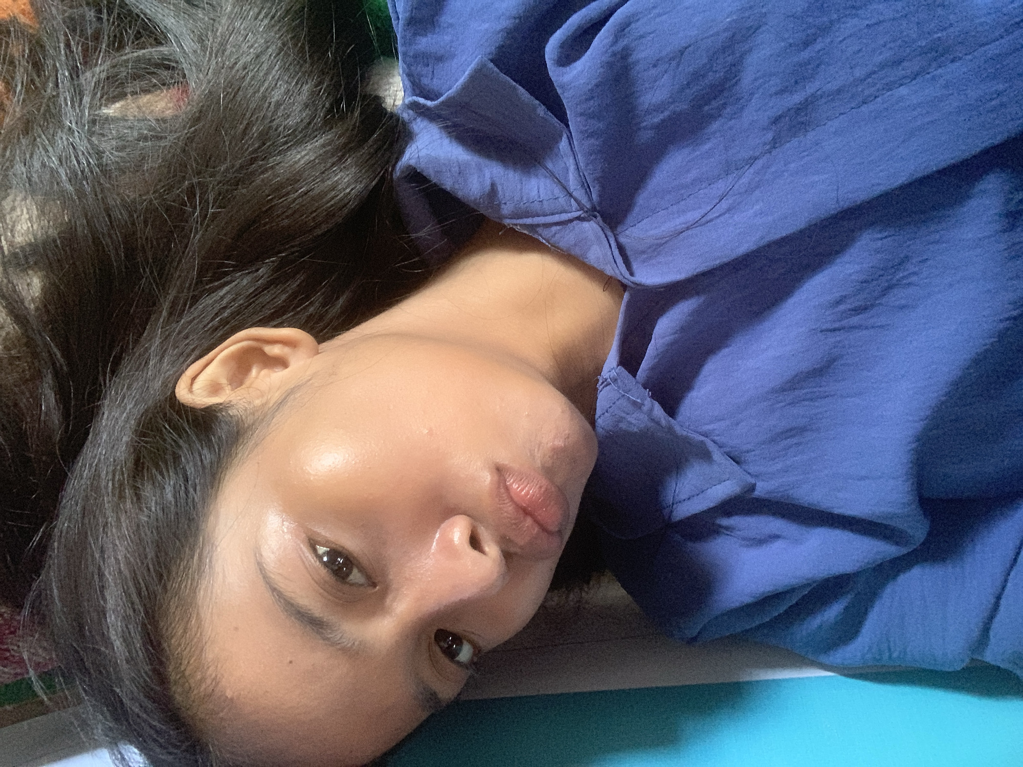 A close-up portrait shows a young woman lying down and looking directly at the camera, with the location context being Sungai Meriam, Indonesia. Her head is tilted, suggesting she is resting on her side or back. She has long, dark hair that spreads out behind her head on the left side of the frame. Her skin has a warm tone and appears smooth and somewhat luminous, particularly on her cheeks and the bridge of her nose. She has dark, expressive eyes and thick, dark eyebrows. Her lips are slightly parted and form a subtle pout, conveying a playful or coy expression.
She is wearing a blue garment, possibly a shirt or dress, made of a lightweight, textured fabric that shows natural creases and folds. Parts of the fabric are visible on the right side of the frame and around her neck area, displaying some stitching. Her right ear is partially visible near her hair on the upper left. The background is largely indistinct, featuring a darker, possibly patterned surface behind her hair and a lighter, likely turquoise-colored surface at the bottom right. The lighting is soft and even, highlighting her facial features without harsh shadows. No text is visible in the image. The scene suggests a casual, intimate moment captured indoors.