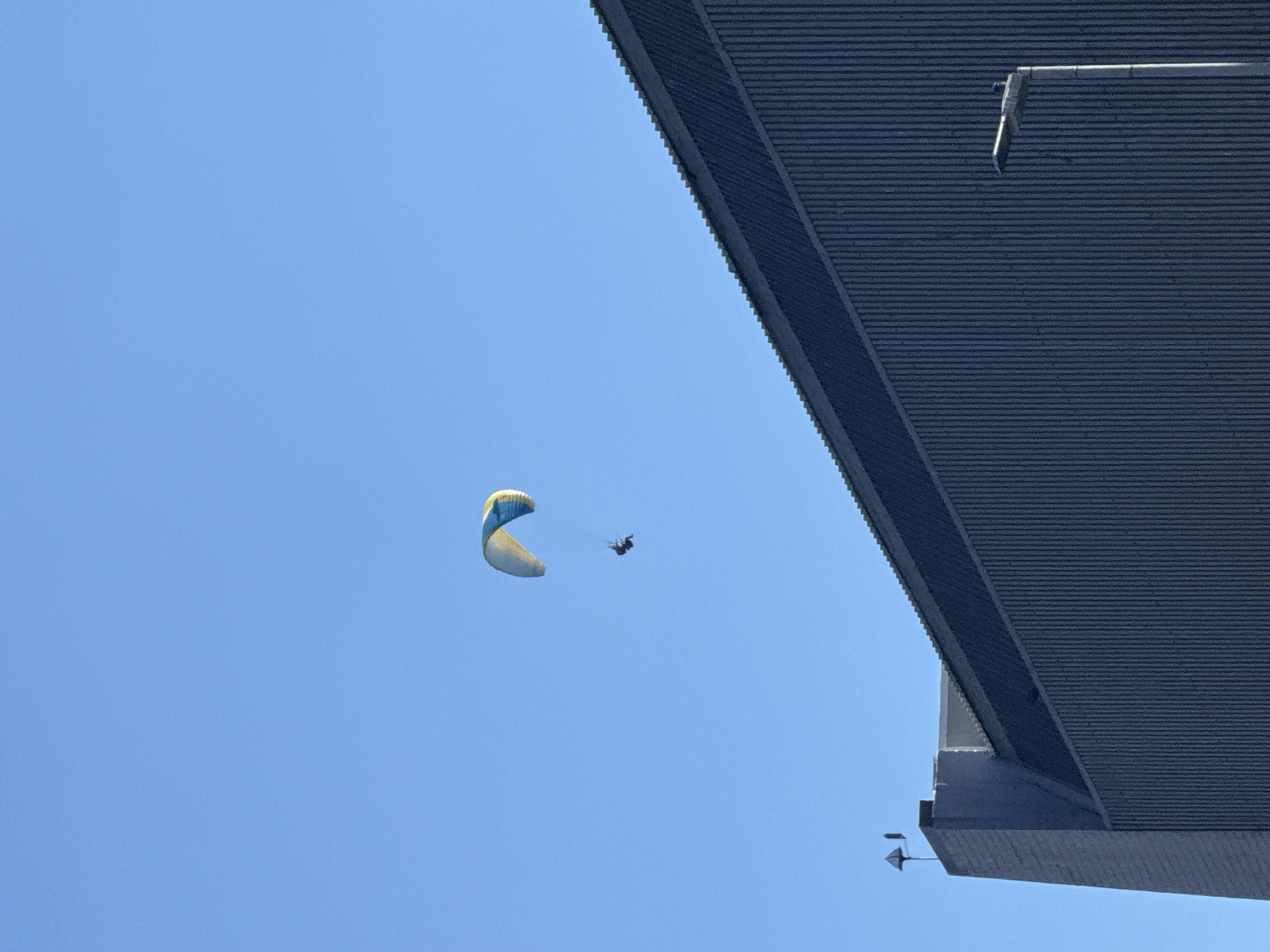 A powered paraglider flies near an unidentified building in Cape Town, South Africa. The building, seen in the upper right corner of the frame, has a corrugated metal roof and appears to be a large structure. The paraglider is light blue and white, with a visible pilot wearing dark clothing. The sky is clear and blue.