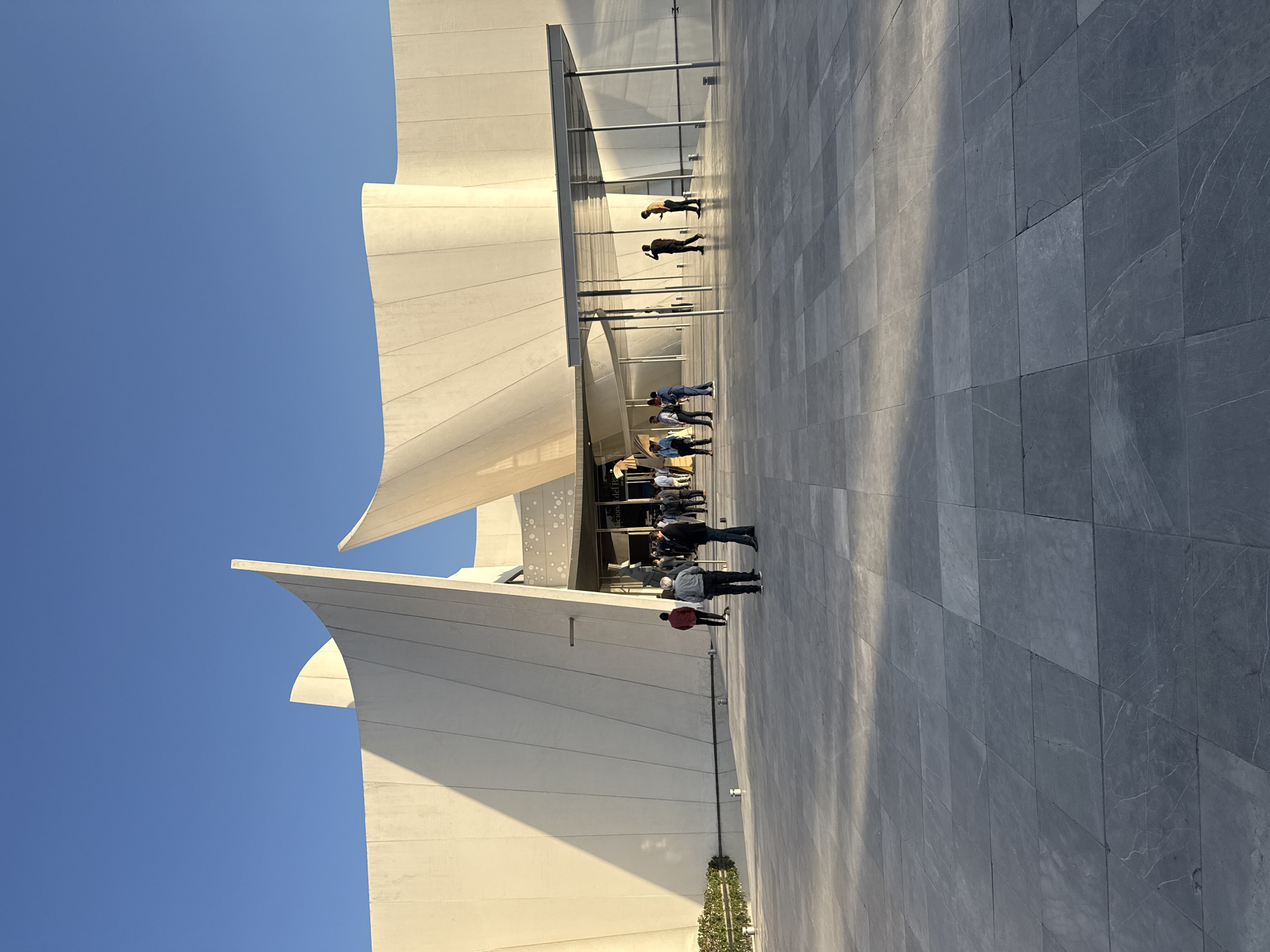 The image displays an outdoor scene in front of a striking modern building under a clear blue sky, likely in San Bernardino Tlaxcalancingo, Mexico. The architecture features prominent, undulating white concrete or stone panels that create a dynamic, sculptural facade, reminiscent of sails or abstract forms. These curved elements rise towards the sky, contrasting sharply with the bright blue.
In the foreground and midground, a wide, grey-tiled plaza extends, reflecting sunlight and casting long, distinct shadows from the people present. This suggests either early morning or late afternoon on a very sunny day. Several individuals and small groups are seen walking or standing on this plaza, congregating near the building's large glass entrance. They are dressed in a variety of casual to semi-casual clothing. Some people are standing closer to the building, while others are further out on the plaza.
A small, green, manicured shrub or plant is visible near the base of the building on the left side. The tiled ground features visible grout lines and some natural veining within the grey tiles, creating a textured surface. The overall impression is one of modern design, open space, and bright natural light. There is some indistinct text visible on the building's glass entrance, partially reading "nacional del P" but the full text is not decipherable.