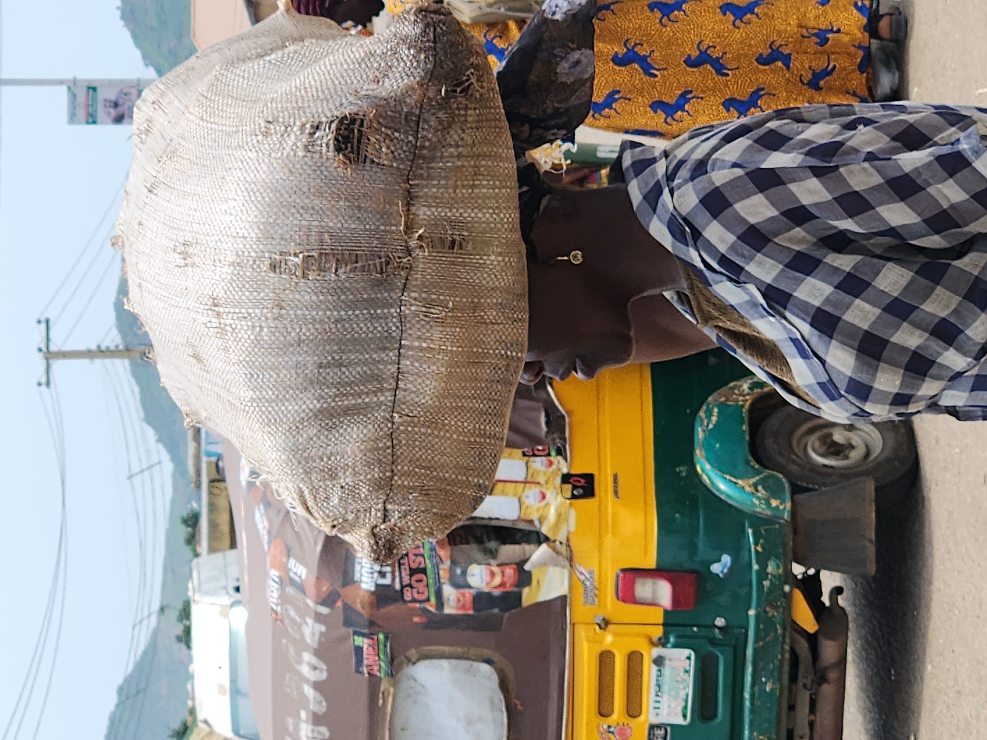 In Kabong, Nigeria, a woman is seen from the side carrying a large, woven sack on her head. She wears a checkered shirt and an earring. Behind her, a yellow and green auto-rickshaw is visible, adorned with various stickers and advertisements. The background shows a street scene with more vehicles and buildings under a hazy sky.