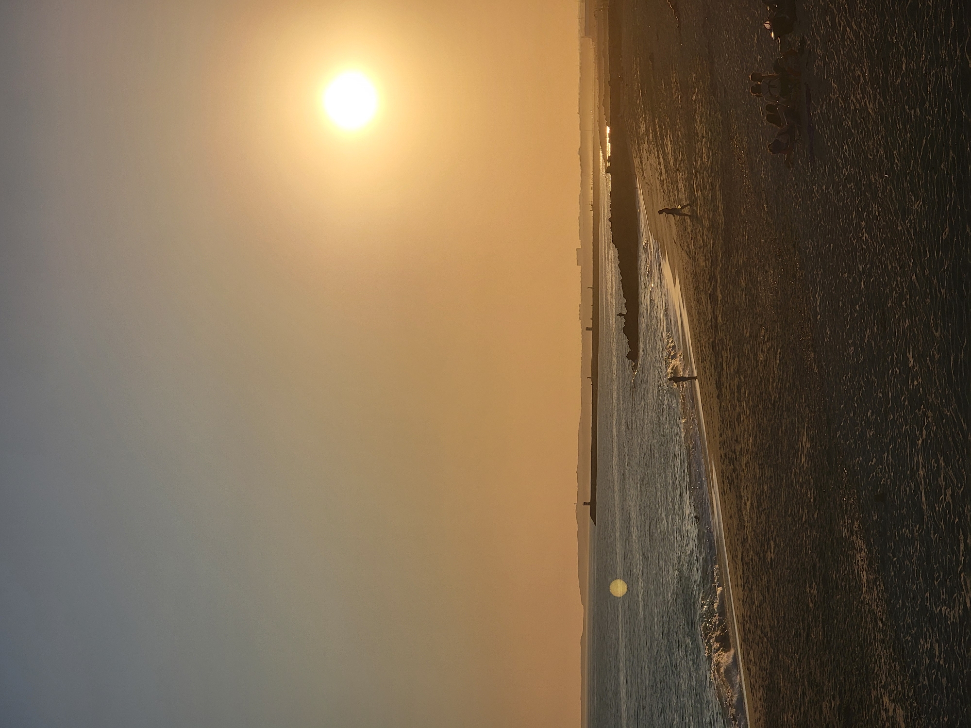 This is a landscape photograph of a beach at sunset in Quarteira, Portugal. The sun is low in the sky, casting a warm, golden glow over the scene and creating a hazy atmosphere. The ocean meets the sandy shore with gentle waves.
Several people are visible on the beach. In the foreground, a lone figure walks along the waterline. Further back, a group of people are gathered, appearing to be relaxing or socializing. Their presence suggests a pleasant, leisurely atmosphere.
The sand is textured and appears to be the primary ground cover, with some patches of wet sand near the water's edge. A pier or breakwater extends into the ocean, providing a linear element in the mid-ground. The overall impression is one of a tranquil end to a summer day, with the warm light and the presence of people enjoying the coastal environment. The time of day is clearly sunset due to the low position of the sun and the warm, diffused light. The weather appears calm and clear, suitable for a beach outing.