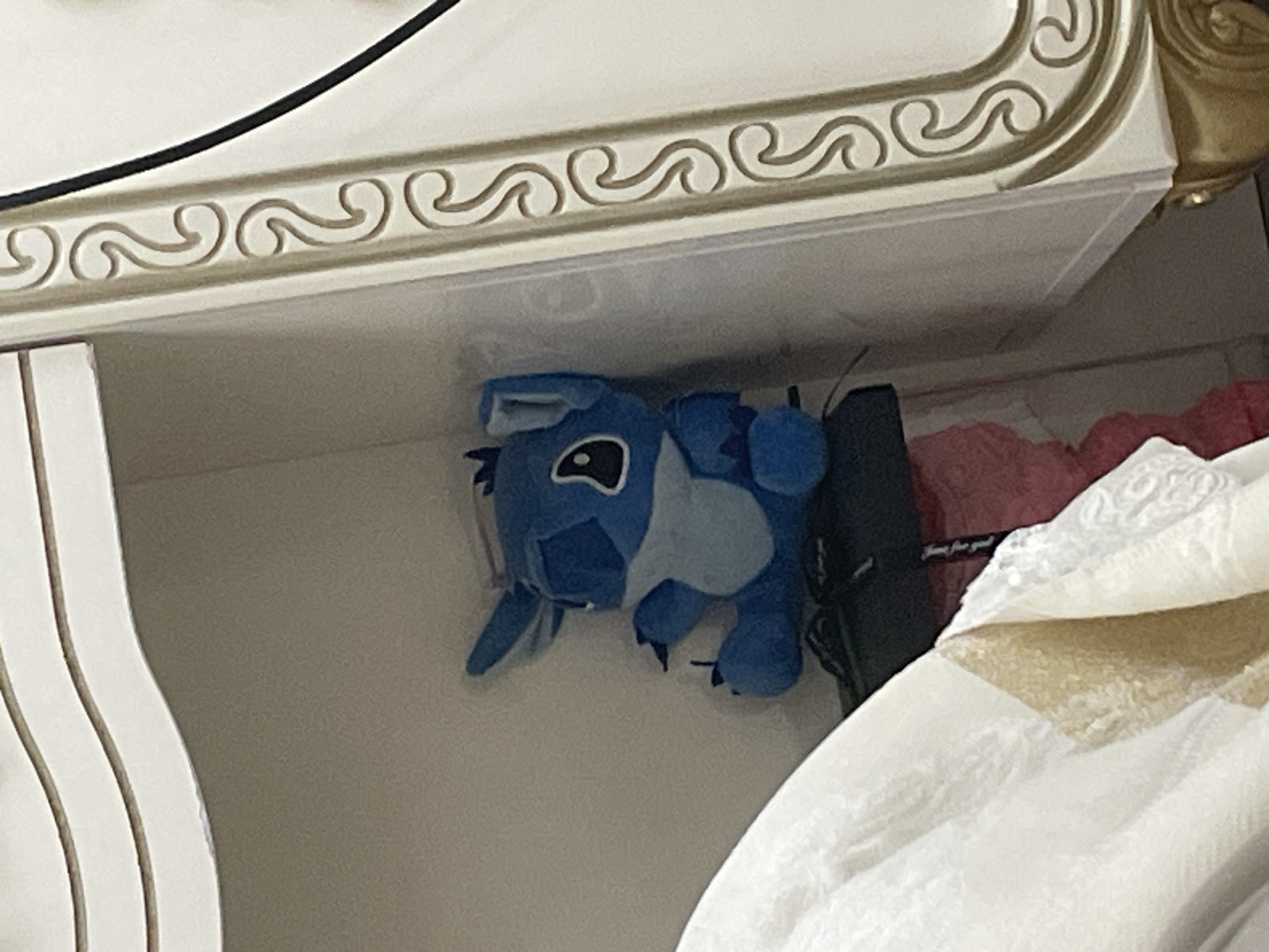 This close-up indoor photograph, taken in Ihtiman, Bulgaria, captures a blue plush toy resembling Disney's Stitch, positioned within a shelf or a recessed area. The plush toy lies on its back or side, facing the viewer, with its prominent black eyes and large ears clearly visible.
Above the toy, a decorative white trim with a repeating wave-like pattern is visible, bordered by a thin gold line. This trim appears to be part of a piece of ornate furniture, possibly a bed frame or a large cabinet. A black cable runs across the top left corner of the image, partially obscuring the trim.
To the left of the plush toy, a white, vertically ridged panel can be seen, which might be part of furniture or a wall. To the right of the Stitch toy, a black object, possibly a bag or case, is tucked next to it. Faint white text is visible on this black object, reading "for you" and possibly "Time for you!" or "Love for you!" preceding it. Below and to the right of the black object, a white, textured fabric, resembling bedding or a blanket, is present, with a hint of red fabric behind it.
The scene appears to be well-lit, suggesting daytime or an adequately illuminated indoor space. There is no direct activity occurring; the image presents a static arrangement of objects in what appears to be a personal living space.