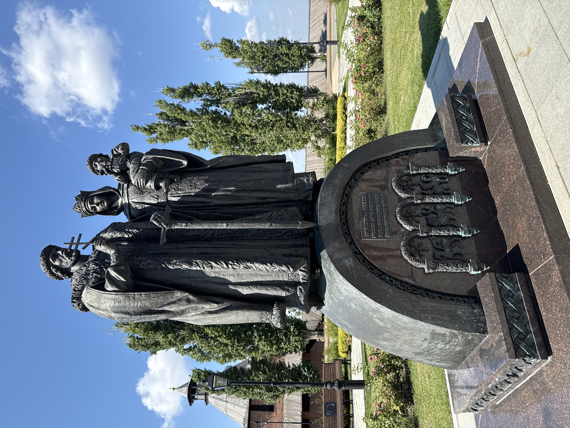 The image shows a bronze monument depicting a man, a woman, and a child on a granite pedestal. The man is on the left, wearing a fur-trimmed robe and holding a cross. The woman, to his right, wears a crown and is holding the child. The child is looking to the left. Behind the monument, there is a wooden building with a tower, green trees, rose bushes, and a blue sky with white clouds. The monument is located in Nizhny Novgorod, Russia, in front of the Nizhny Novgorod Kremlin. The inscription on the monument is in Russian and commemorates Saint Daniel of Moscow.