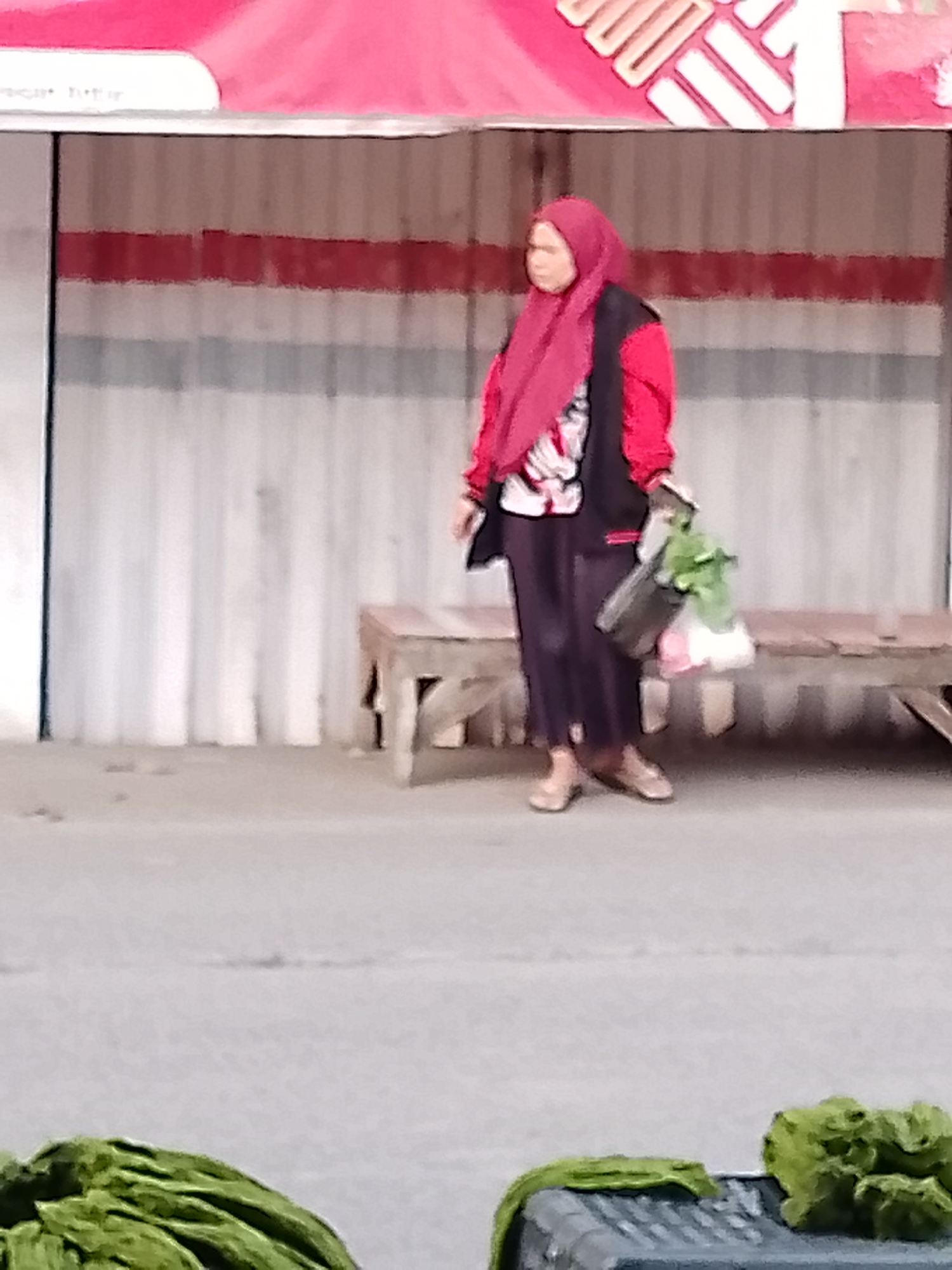A woman wearing a red hijab and a red and black jacket stands on a sidewalk in Cikalongkulon, Indonesia. She is holding a green bag with produce in her right hand and another bag in her left hand. In front of her is a low wooden bench. The background is a corrugated metal wall with red and white stripes. In the foreground, there is a grey trash can with green leafy vegetables.