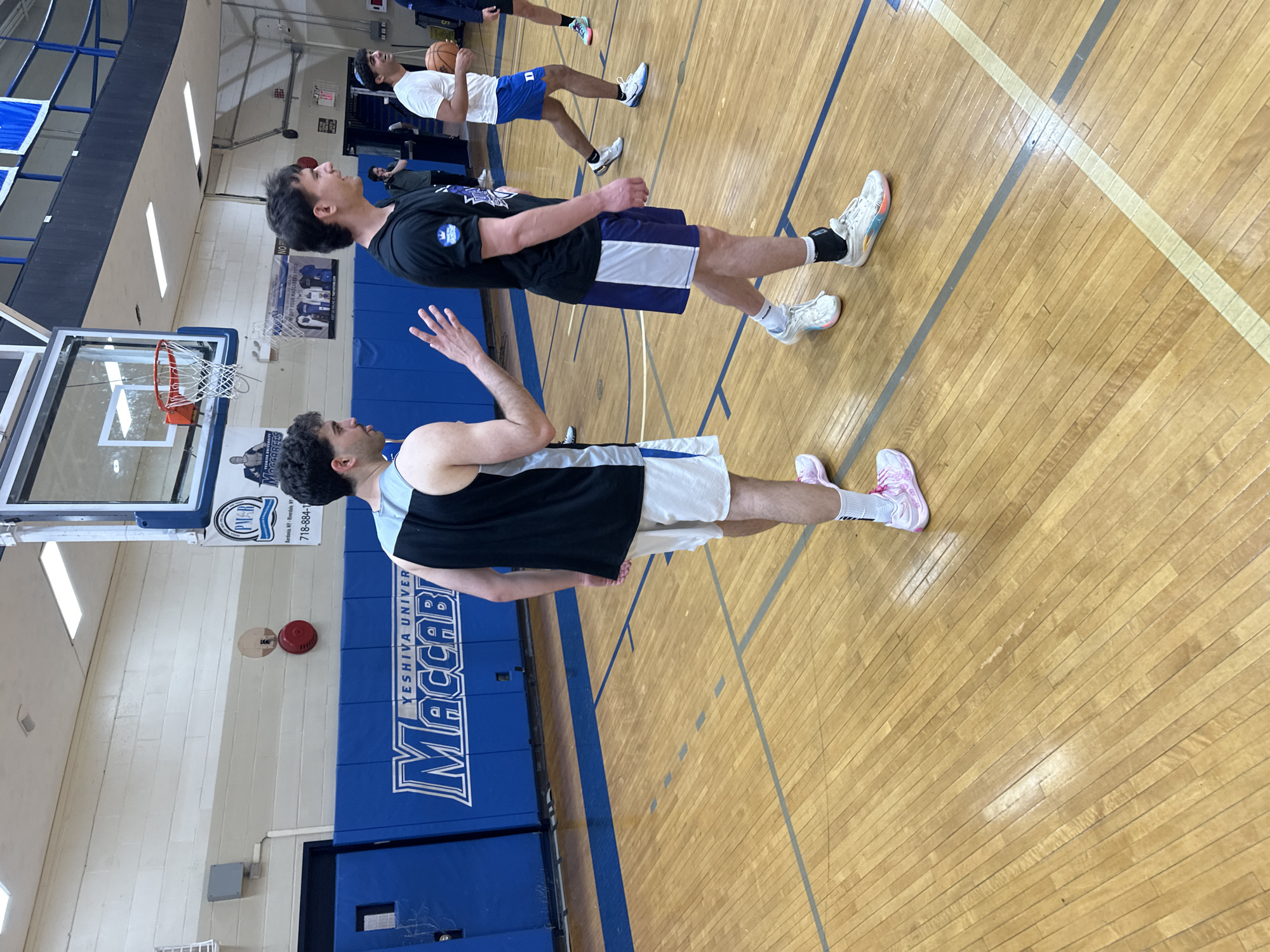 An indoor basketball court is depicted, with several young men, likely basketball players, on the wooden floor. The image is taken from a low angle, looking across the court. In the foreground, two men stand side-by-side. One on the right wears a black t-shirt, blue and white shorts, and white sneakers with colorful accents. The other, on the left, wears a black and light blue tank top, white shorts, and pink and white sneakers. Their expressions are neutral, appearing to be observing or waiting. Further back, another player in a white t-shirt and blue shorts holds a basketball.
The setting is a gymnasium with light wooden floorboards marked with blue and yellow court lines. Blue padded walls line the side of the court, featuring prominent white text that reads "YESHIVA UNIVER MACCABI" and "MACCABEES". Above the padding, a white wall has a sign with "PM" and location information "Bardonia, NY - Riverdale NY" along with a partial phone number "718-884-1". Another sign partially visible says "YOUR MACCABEES PRICE". A red fire alarm is mounted on the wall above the blue padding. A basketball hoop with a clear backboard and orange rim is visible on the left side of the frame. The lighting suggests an indoor daytime setting.