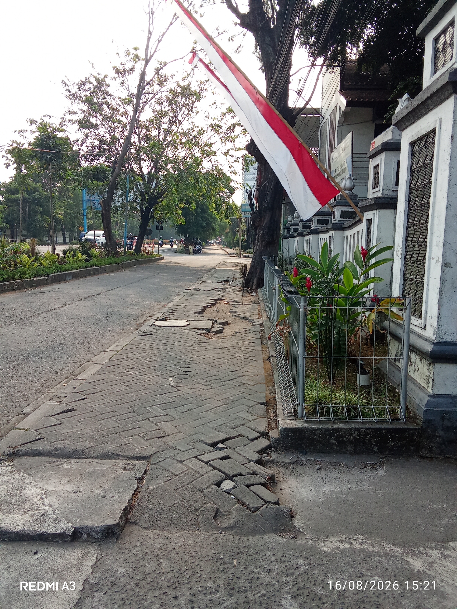 A street scene in Surakarta, Indonesia shows a paved road on the left with a damaged cobblestone sidewalk on the right. A red and white Indonesian flag flies from a bamboo pole attached to a grey and white building. Green trees and plants line the street, with some vehicles and pedestrians visible in the background. The sky is overcast and bright. The timestamp "16/08/2026 15:21" and "REDMI A3" are visible in the bottom right and left corners respectively.