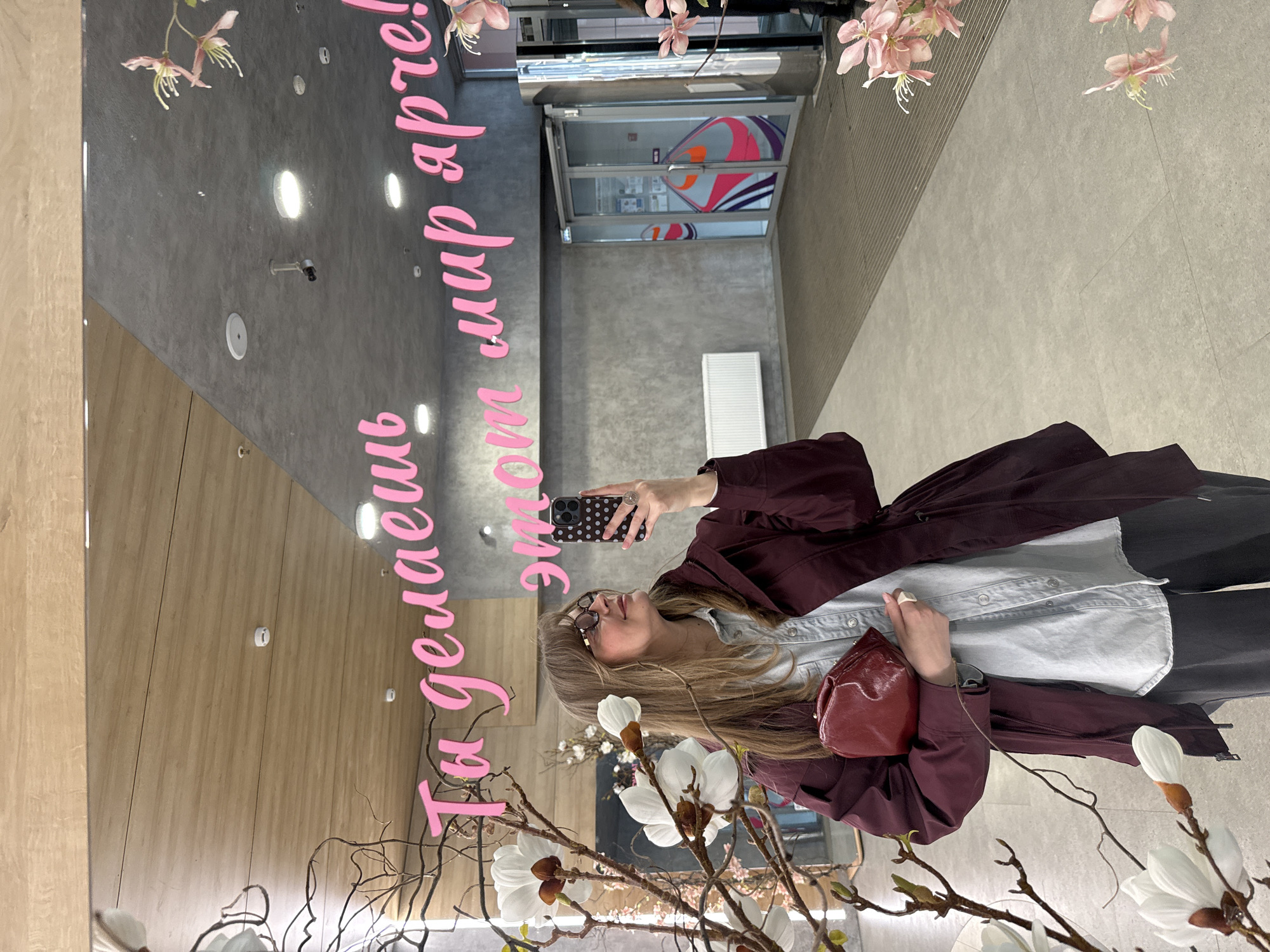 A blonde woman is taking a selfie in a mirror, holding a phone with a black and white dotted case in her left hand, wearing glasses and a maroon jacket. She holds a small red purse with her right hand. The mirror reflects a commercial space, featuring light-colored wood paneling, a gray ceiling with recessed lighting, and decorations of artificial magnolia blossoms. A pink script reads "Ты делаешь этот мир ярче!" (You make this world brighter!) in Russian. The location appears to be a public space, likely a mall or shopping center, in Izhevsk, Russia, based on the Russian script.