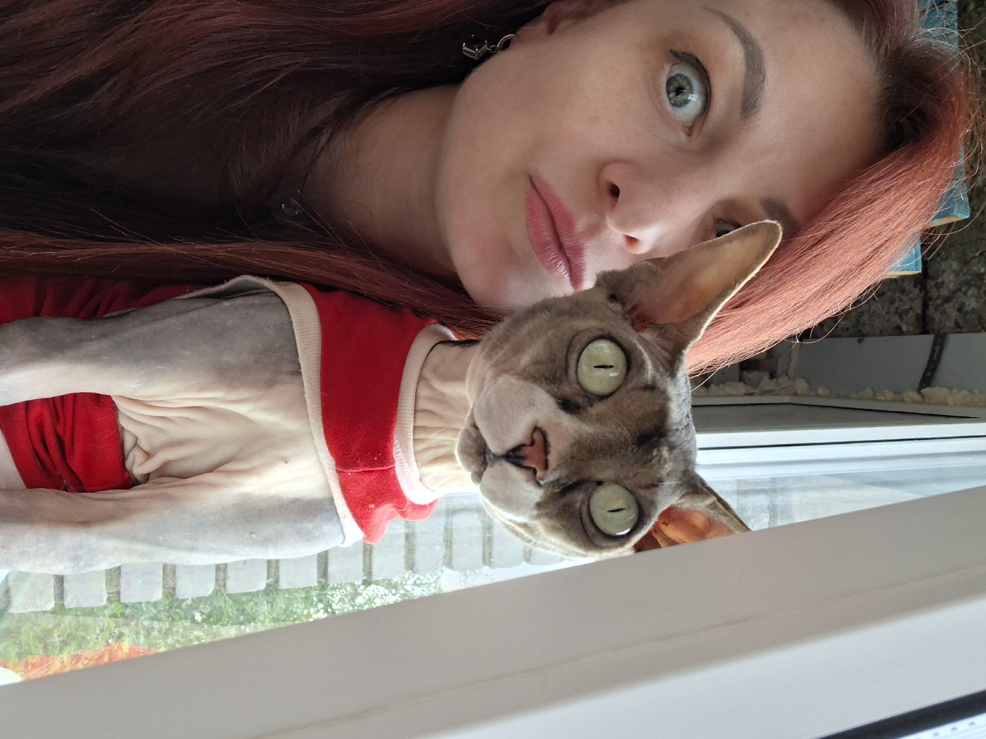 This is a close-up, rotated selfie featuring a woman and a Sphynx cat. The woman has long, reddish-brown hair and is looking at the camera with wide, blue-green eyes. She has light skin and her lips are a soft pink. She is wearing a red garment with a light-colored trim, which partially covers the cat.
The cat is a Sphynx with distinctive greyish skin and large, pale green eyes with slit pupils. It is wearing a red garment, likely a sweater or shirt, with a white neckline and trim. The cat's face is turned towards the camera, with its ears perked up.
The scene appears to be indoors, near a window. Through the window, there is a blurred view of green foliage, suggesting it is daytime and possibly spring or summer. The woman and cat are positioned in front of the window, with a white frame partially obscuring the lower part of the image.
The overall mood is intimate and casual, possibly capturing a moment of interaction between the woman and her pet. The lighting suggests it is daytime. There is no visible text in the image. The location context provided is Забалоцкі сельскі Савет, Belarus.