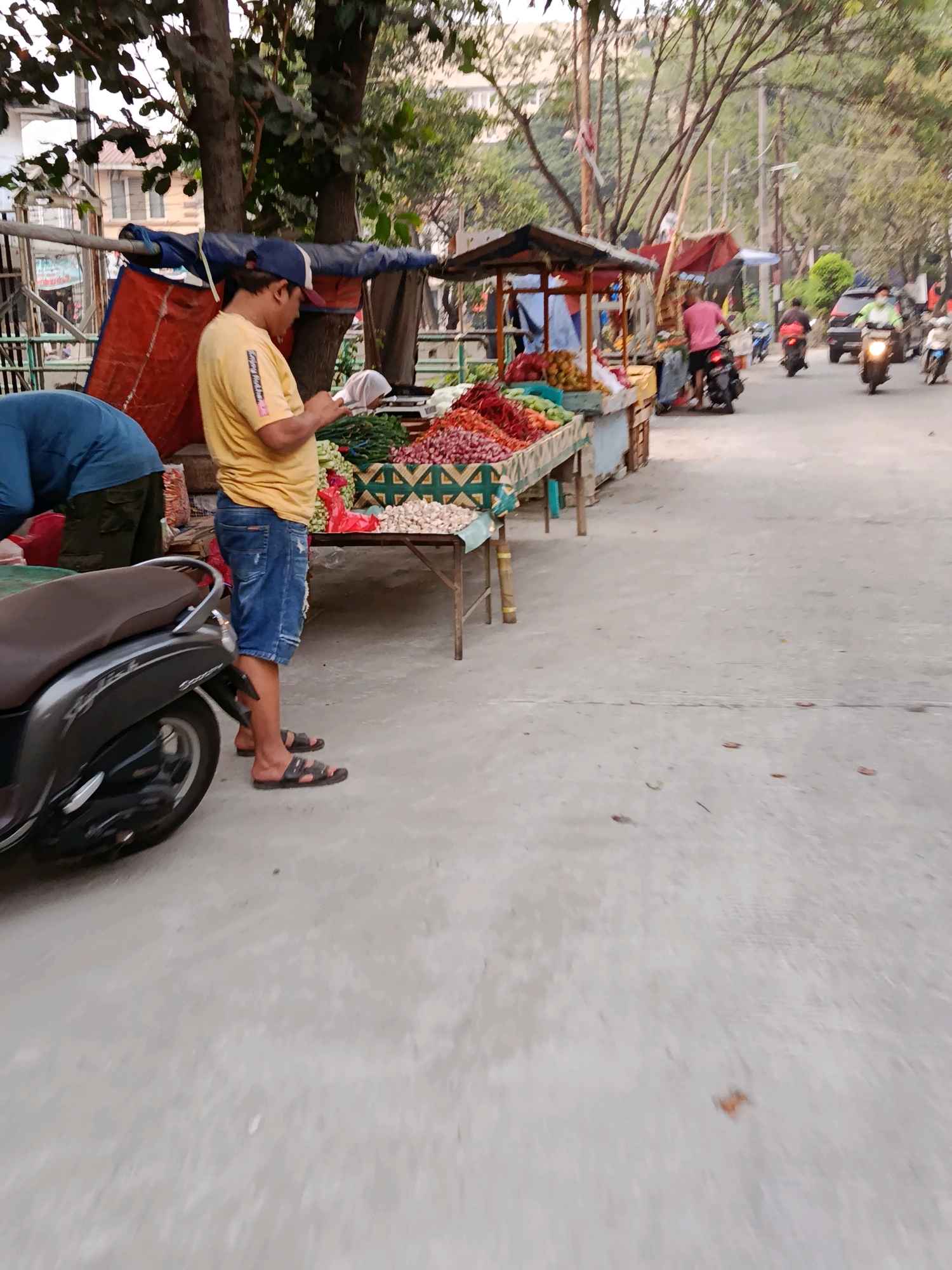A man in a yellow t-shirt and denim shorts stands on a paved street in Bekasi, Indonesia, looking at his phone. He is positioned between a parked scooter on his left and a market stall on his right. The market stall displays a variety of fresh produce, including red onions, chilies, garlic, and leafy green vegetables. Behind the stall, a blue tarp covers some items, and beyond that, trees and buildings are visible. Other people and motorbikes can be seen in the background on the street. The overall scene depicts a typical street market environment in an urban setting.