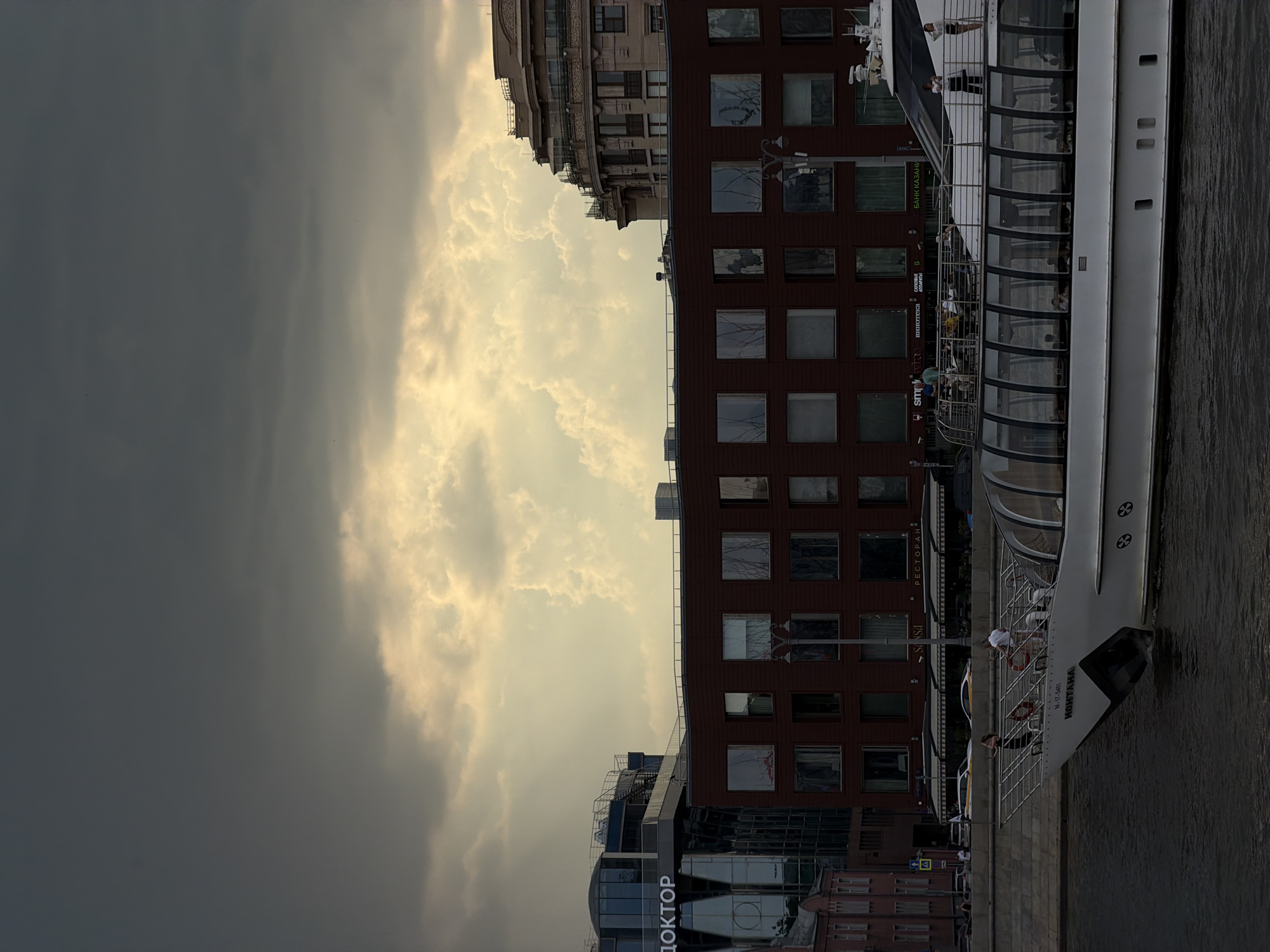 The image depicts a boat, identified as "KOHAHA," docked on a river embankment in Moscow, Russia. The sky is dominated by heavy, dark clouds on the left, contrasting with a section of brighter, sunlit clouds in the center. Alongside the embankment, a modern brown brick building with multiple windows stands next to a glass-fronted building with Cyrillic lettering. People are visible both on the boat and on the embankment. The perspective is unusual, rotated 90 degrees.