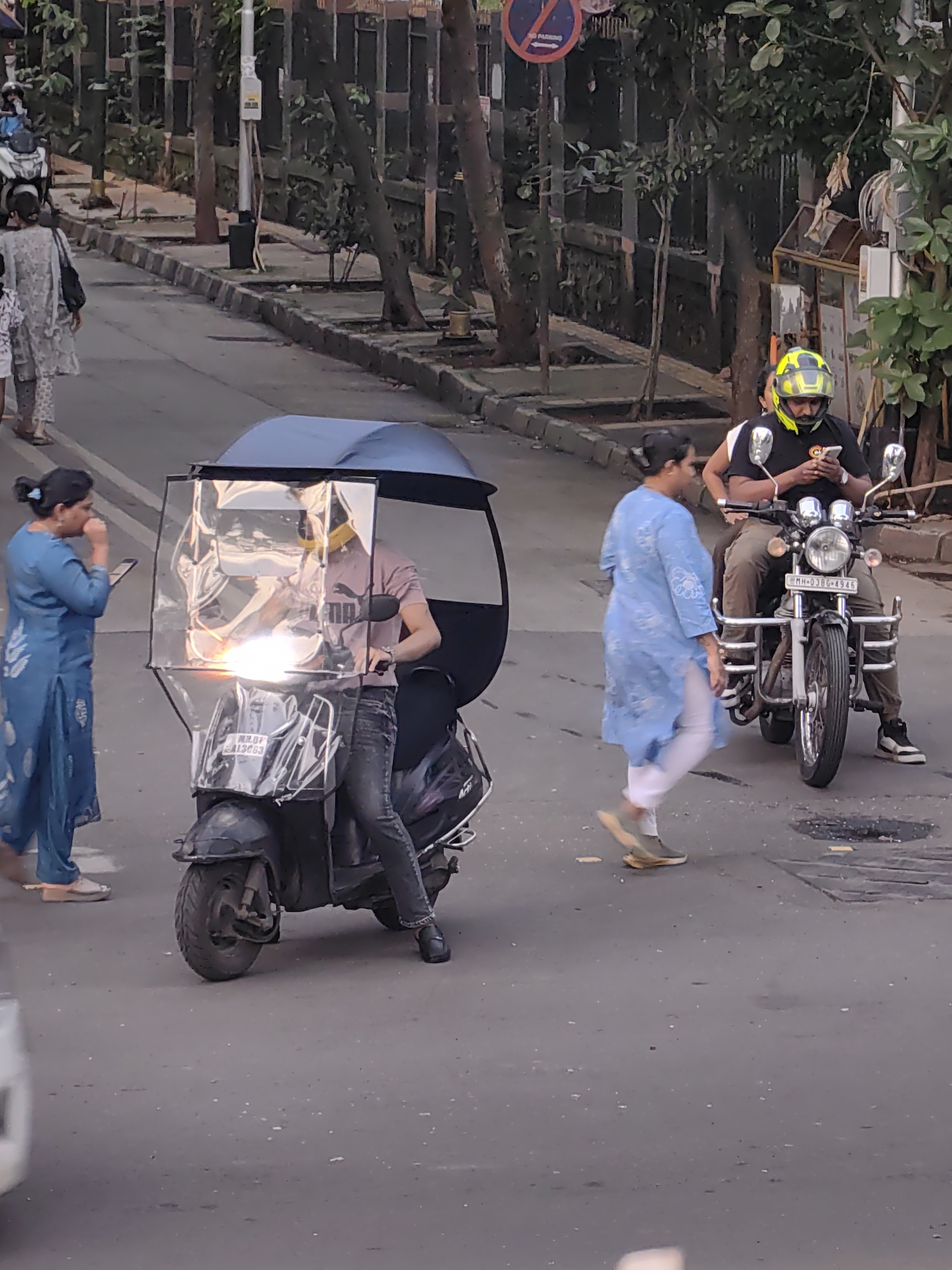 The image captures a street scene in Mumbai, India, likely during the day. The foreground features a scooter with an attached clear plastic shield and a dark blue umbrella, providing cover for the rider. The rider, wearing a pink t-shirt and dark jeans, is looking forward. To the left, a woman in a blue kurta and white pants is standing, holding a phone and appearing to talk. To the right, a man wearing a yellow helmet, black t-shirt, and brown pants is riding a motorcycle and looking at his phone. Another person, partially visible behind him, is also on the motorcycle. The background shows trees, a sidewalk, and a street with other vehicles suggested by blurred shapes. A "No Parking" sign is visible on a pole in the background. The road surface is asphalt with some water or oil stains and scattered debris.