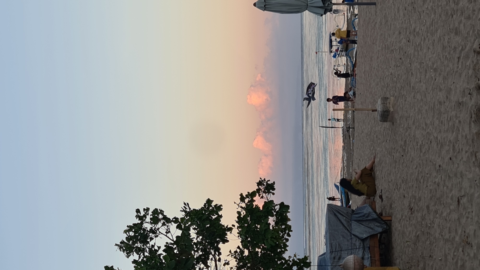 The image depicts a beach scene in Denpasar, Indonesia during twilight. The sky transitions from a light blue at the top to a soft pink and yellow hue near the horizon. Fluffy pink clouds are visible in the distance. The ocean is calm with gentle waves, reflecting the colors of the sky. On the sandy beach, several people are scattered. In the foreground, a woman with long dark hair is sitting on the sand, facing away from the camera, wearing a yellow top and brown pants. To her left, a wooden structure is covered with a grey tarp. Further down the beach, a kite shaped like a bird is flying in the air. Several small boats are docked on the shore. Groups of people, including children and adults, are standing and interacting near the boats. A large closed umbrella is visible on the right side of the image. The overall atmosphere is serene and peaceful, characteristic of a sunset on a tropical beach.