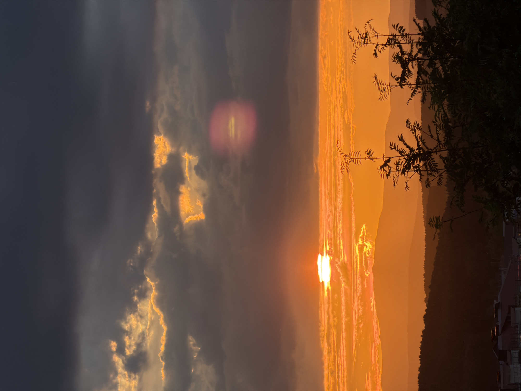 This is a vertical, high-angle, full shot of a sunset. The sun is a bright orb positioned on the right side of the image, with rays of light emanating from it, creating a vibrant orange glow that illuminates the clouds. The sky is a dramatic mix of dark gray clouds on the left and brightly lit, fiery orange clouds on the right. A faint, circular lens flare is visible in the dark gray portion of the sky. Silhouetted against the sunset are distant, layered hills, covered in trees, and in the far right foreground, a dark silhouette of a tree branch with sparse leaves, and part of a building with illuminated windows. The overall impression is one of a dramatic and beautiful natural phenomenon. The time of day is clearly sunset, given the intense colors and the position of the sun. The weather appears to be dynamic, with both dark clouds and illuminated ones, suggesting a transition or an impactful display of atmospheric light. The presence of a building suggests a possible human settlement or developed area within or near the natural landscape. The scene evokes a sense of awe and the power of nature. There is no visible text in the image.
