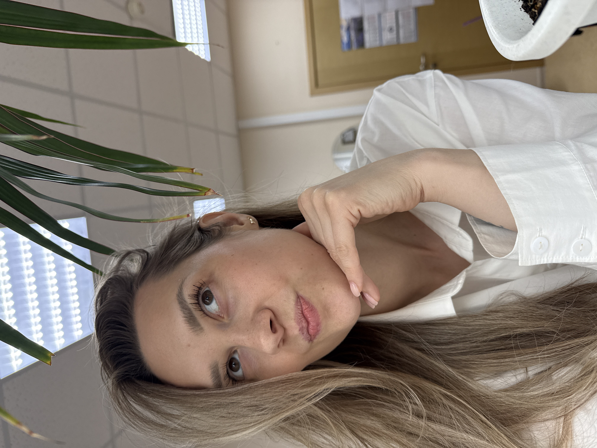 The image shows a woman captured from a tilted, close-up perspective, making it appear as if she is lying down and looking upwards. Her long, light brown hair cascades around her head. She has a calm and contemplative expression, with natural-looking makeup, including mascara, and light pink nail polish on her fingers. Her right hand is gently placed near her chin. She is wearing a white collared shirt with visible buttons.
In the foreground on the left, several long, green leaves of a plant partially frame her face. The background reveals an indoor setting, consistent with an office or commercial building, featuring white ceiling tiles and bright, rectangular fluorescent light fixtures. A white potted plant is visible in the upper right corner, and elements like a wooden door frame or framed object can be seen further back. The lighting is predominantly bright and artificial. The scene takes place in Chaikovsky, Russia. There is no visible text.