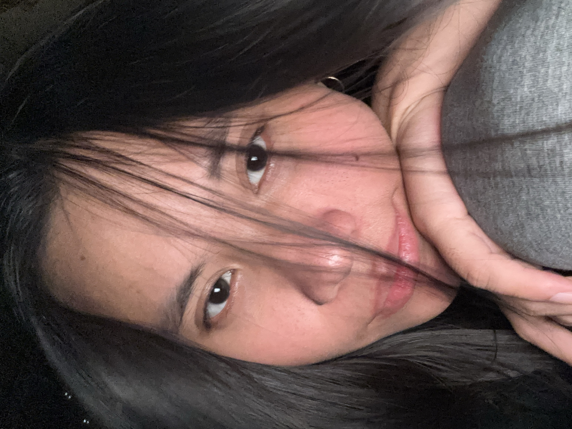 The image is a close-up selfie of a young woman with long, dark hair that partially obscures her face. She is leaning on her arm, which is resting on her leg, wearing a grey garment. Her eyes are open, and her gaze is directed slightly off-camera. Her lips are slightly parted. The background is dark and indistinct, suggesting an indoor setting or nighttime. The overall lighting is soft, with a pinkish hue on her skin, indicating likely artificial light. There are no discernible features to place this image in East Jakarta, Indonesia.