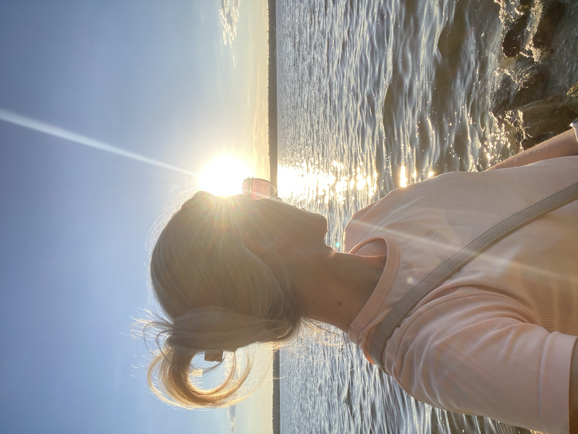 The media file shows a profile view of a person with blonde hair, looking up at the sun. The sun is bright and low in the sky, suggesting it is either sunrise or sunset. The person is wearing a light pink t-shirt and appears to be standing near water, possibly a lake or ocean, as there are waves and rocky shore visible. The overall mood is serene and peaceful, with the warm glow of the sun illuminating the scene. The image is captured in portrait orientation, with the person's head and shoulders in the foreground. The sky is clear and blue, with some wispy clouds. The sunlight creates a lens flare effect, adding to the atmospheric quality of the image. There is no visible text in the media file.