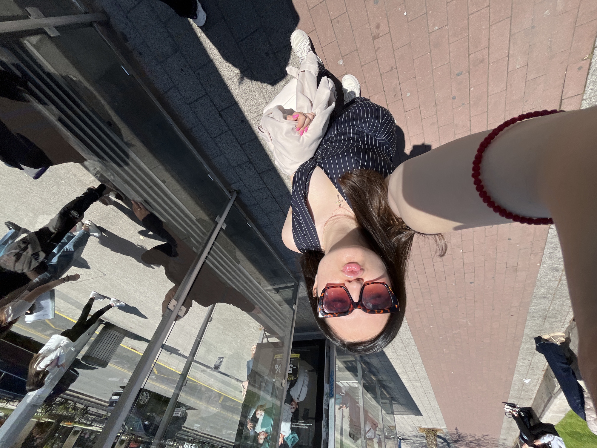 The image presents an outdoor urban scene from an unusual, rotated, high-angle selfie perspective, suggesting the camera is held overhead looking down. In the foreground, a young woman with long dark hair and large brown sunglasses looks up at the camera with a playful pouting expression. She is wearing a striped sleeveless top and a delicate necklace, with a red beaded bracelet visible on her arm.
Beside her, another person is seated on the ground, wearing light-colored clothing and white sneakers, with bright pink nail polish visible on their fingers. The ground features sections of reddish-brown brick paving and grey concrete. Bright sunlight illuminates the scene, casting distinct shadows and indicating it is daytime.
Large reflective glass surfaces, likely from a building, dominate the background, showing distorted reflections of other pedestrians, urban structures, and a car. A metal railing is also visible. Partially legible text on a reflected poster includes "-5%" and parts of a word like "TRA," indicating an advertisement. The scene depicts a casual moment in a bustling city environment, with the provided context identifying the location as Plovdiv, Bulgaria.